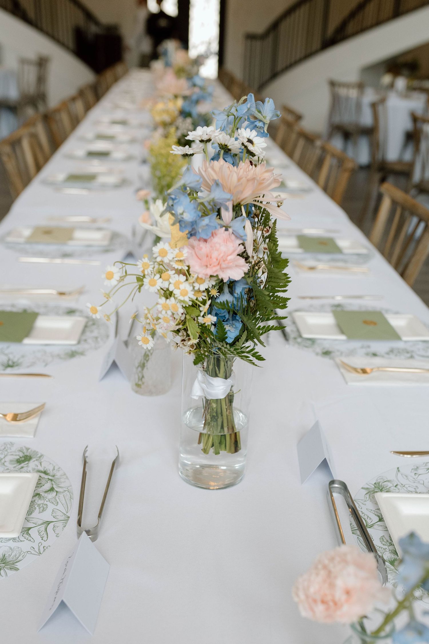 Long elegant wedding reception table with pastel pink and blue floral centerpieces and green botanical place settings.
