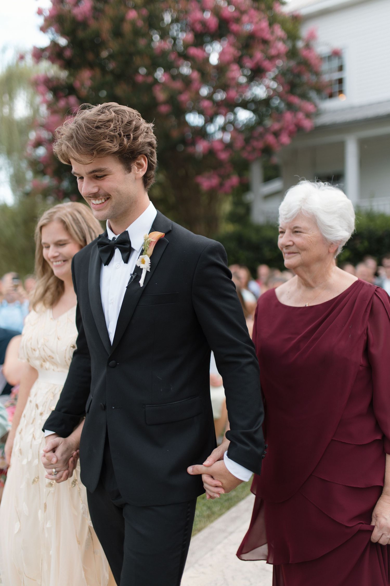 Smiling groom in black tuxedo walks down aisle with mother in burgundy gown and bridesmaid in gold dress.