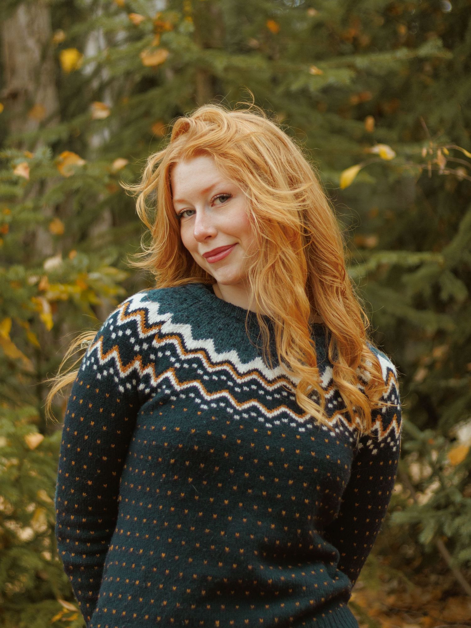 Person wearing a black and white Nordic pattern sweater poses outdoors among autumn foliage.