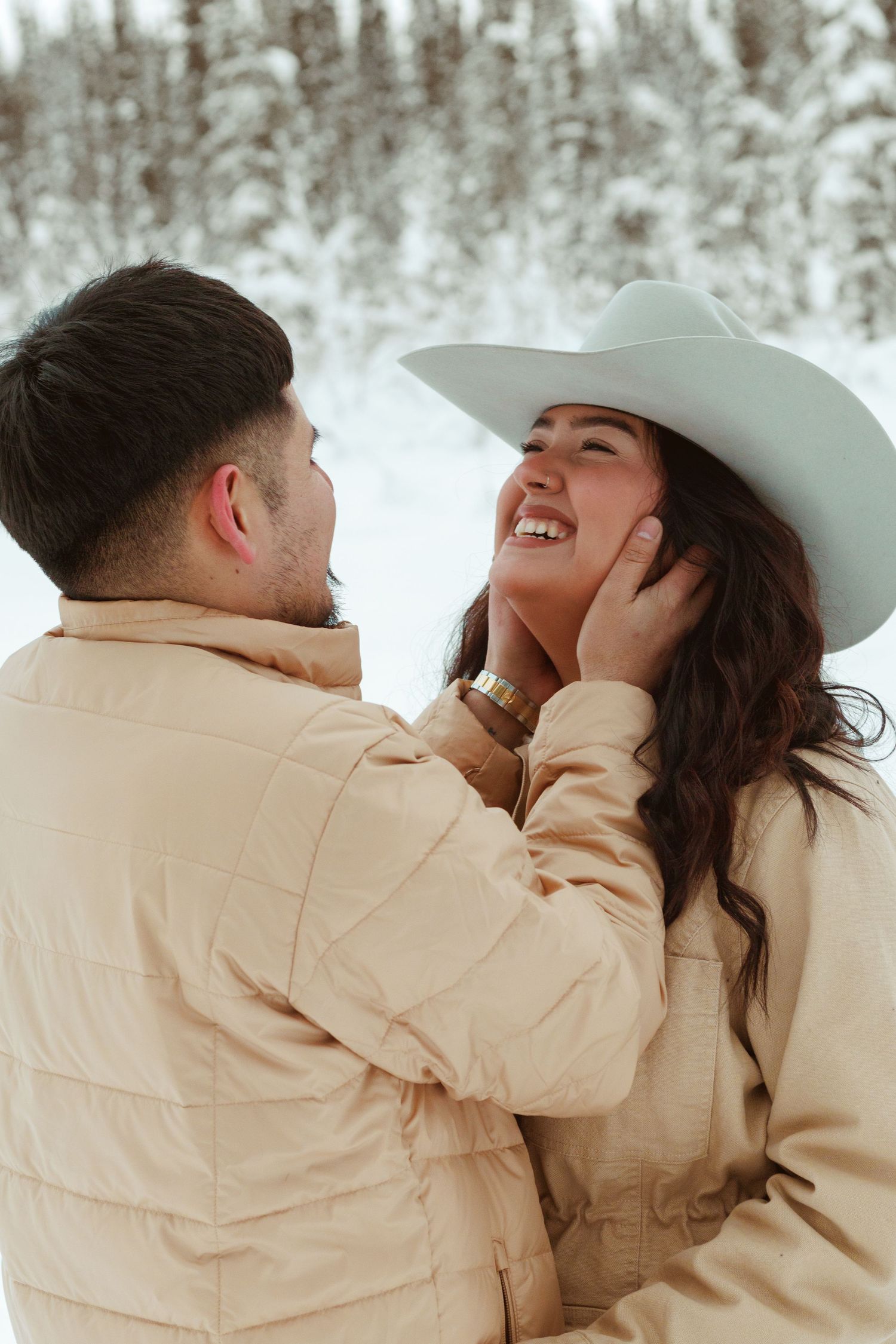 A romantic winter moment between a couple in matching beige coats with a snowy forest backdrop.