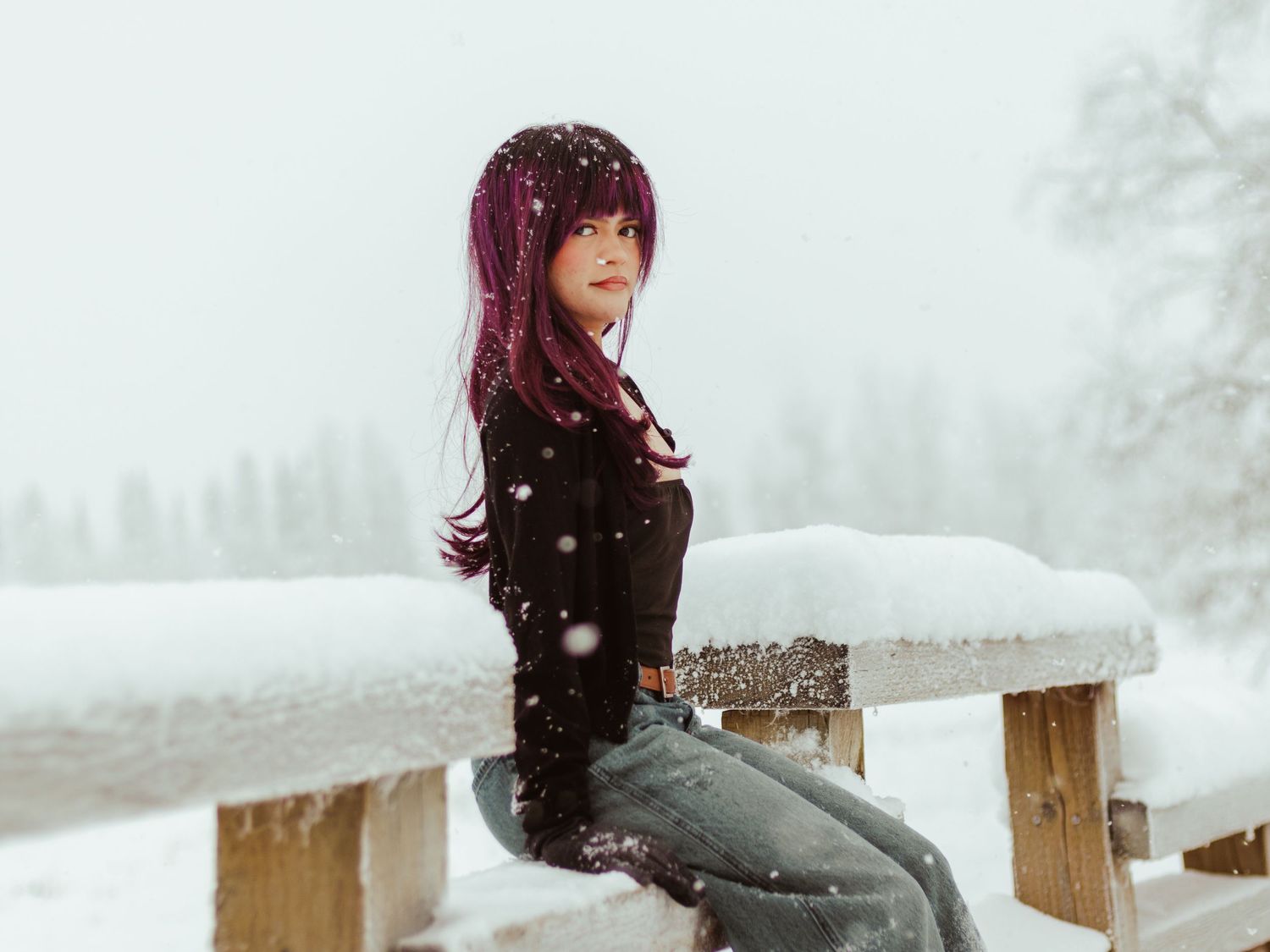 A person with burgundy hair sits on a wooden fence in a snowy winter landscape during snowfall.