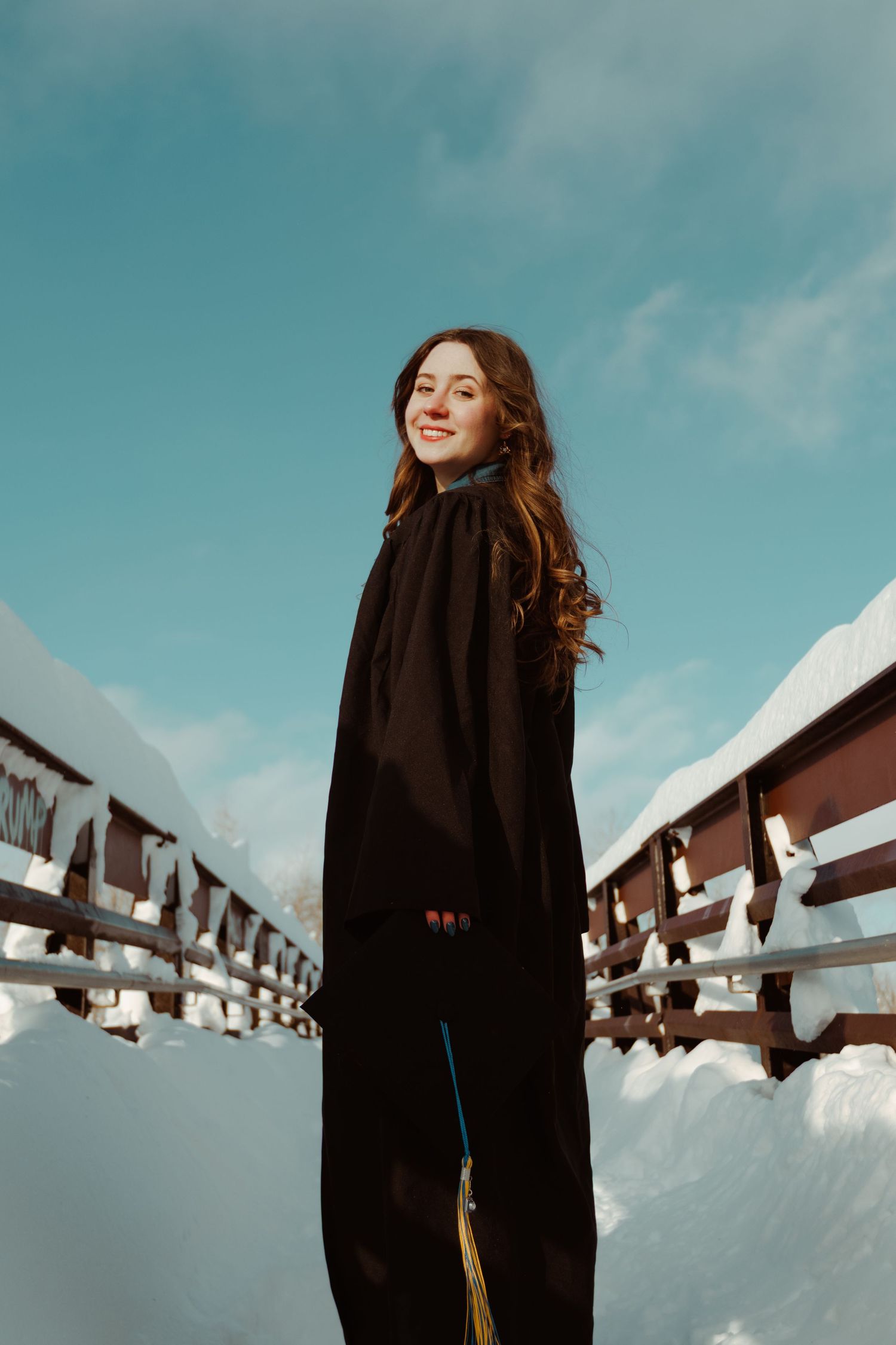 A figure in a black coat stands against a snowy winter landscape with wooden fences and bright blue sky.