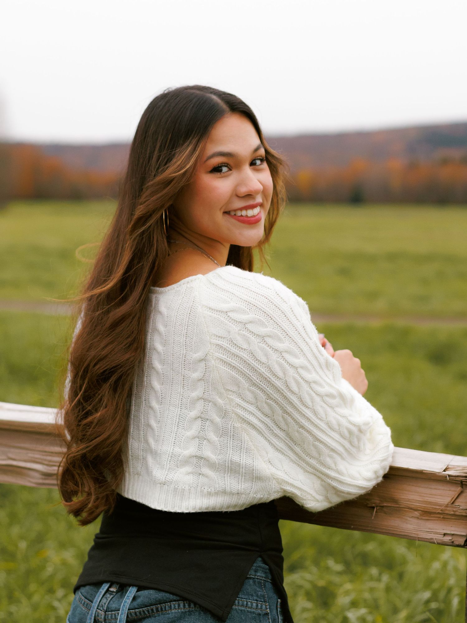 A person in a white cable knit sweater and jeans poses casually against a wooden fence in an autumn field.