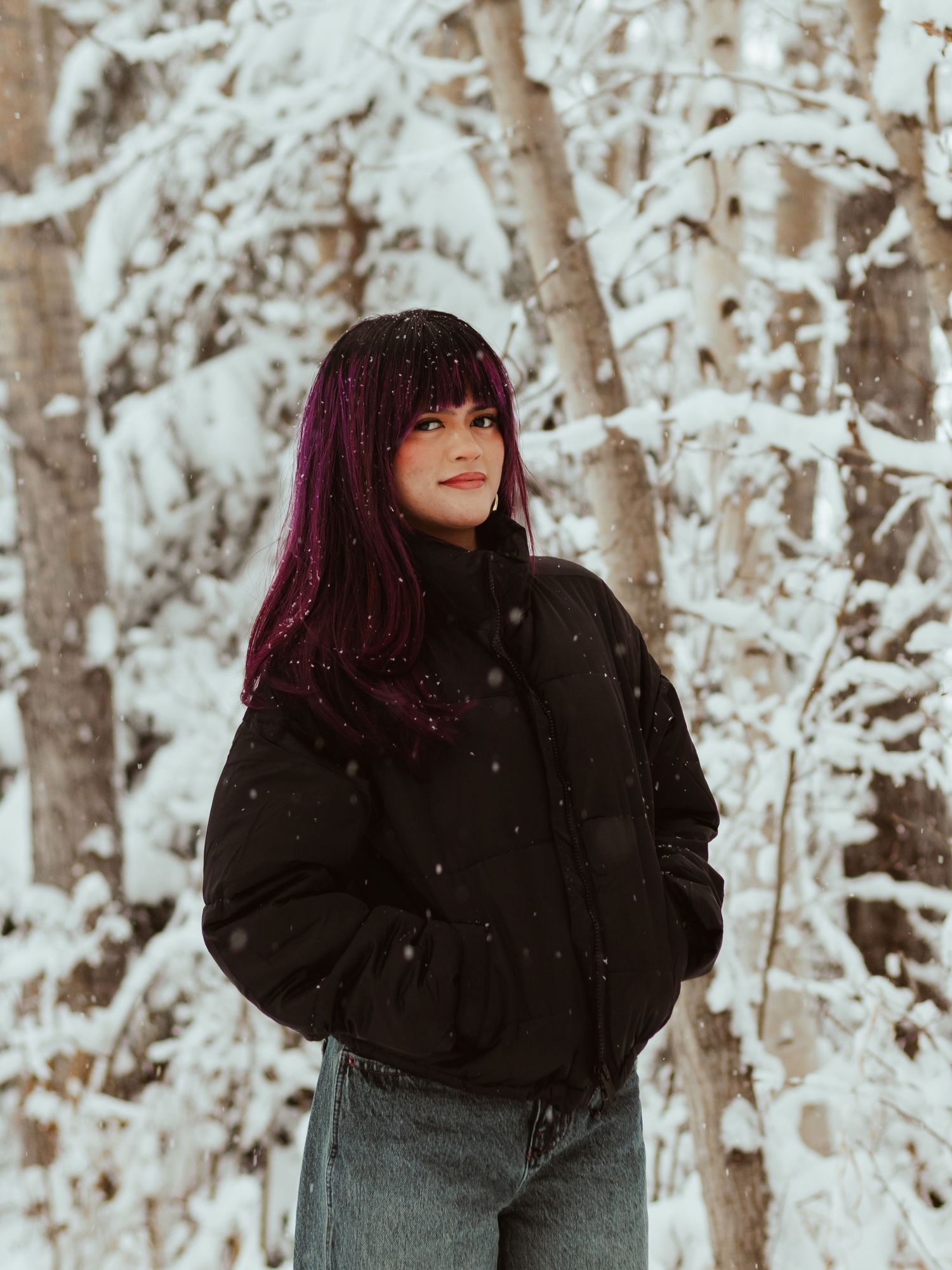 A person in a black winter coat stands in a snowy forest setting with white birch trees in the background.