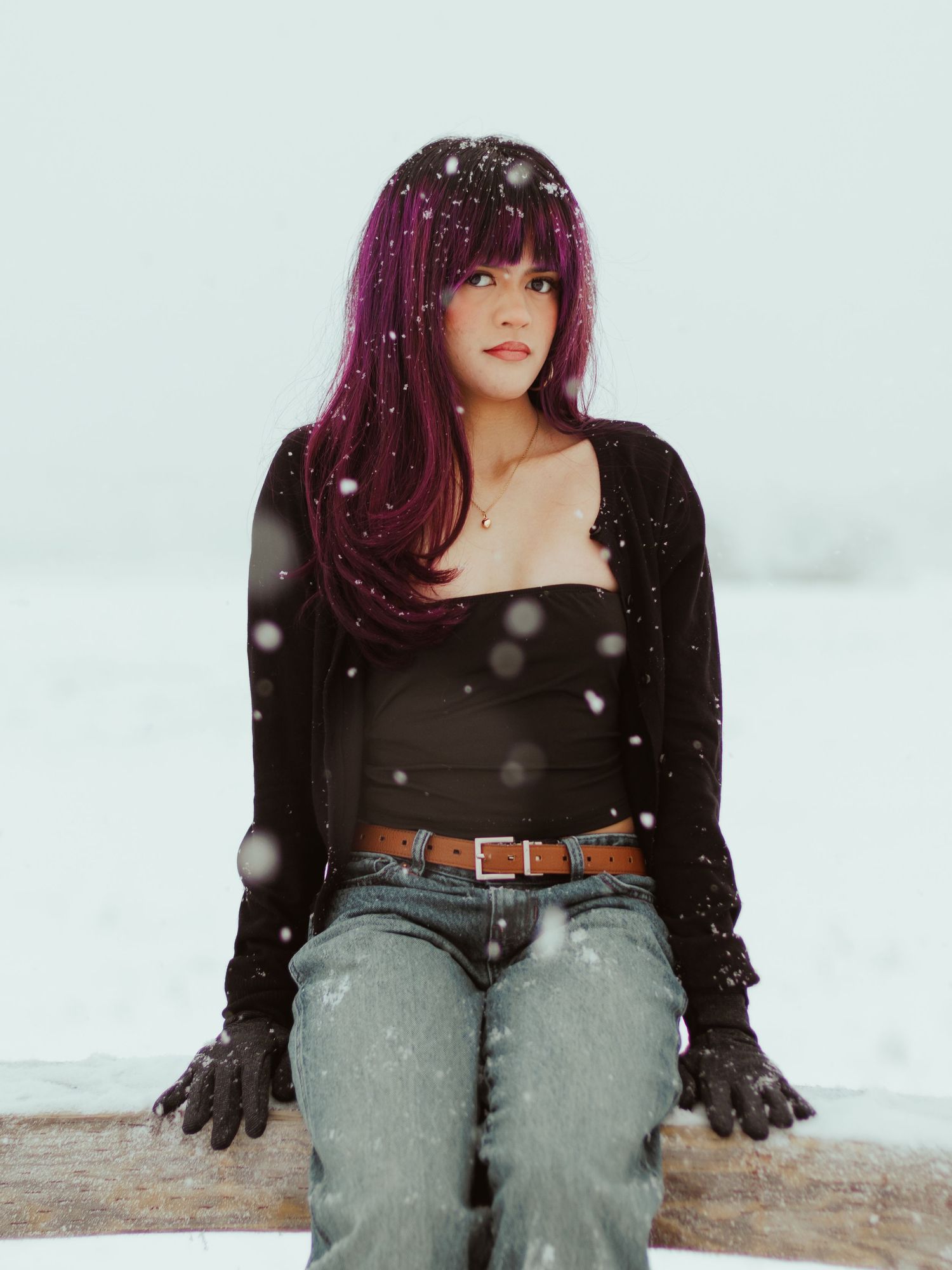 A person with purple hair wearing a black top and jeans sits in snow during a winter photoshoot.