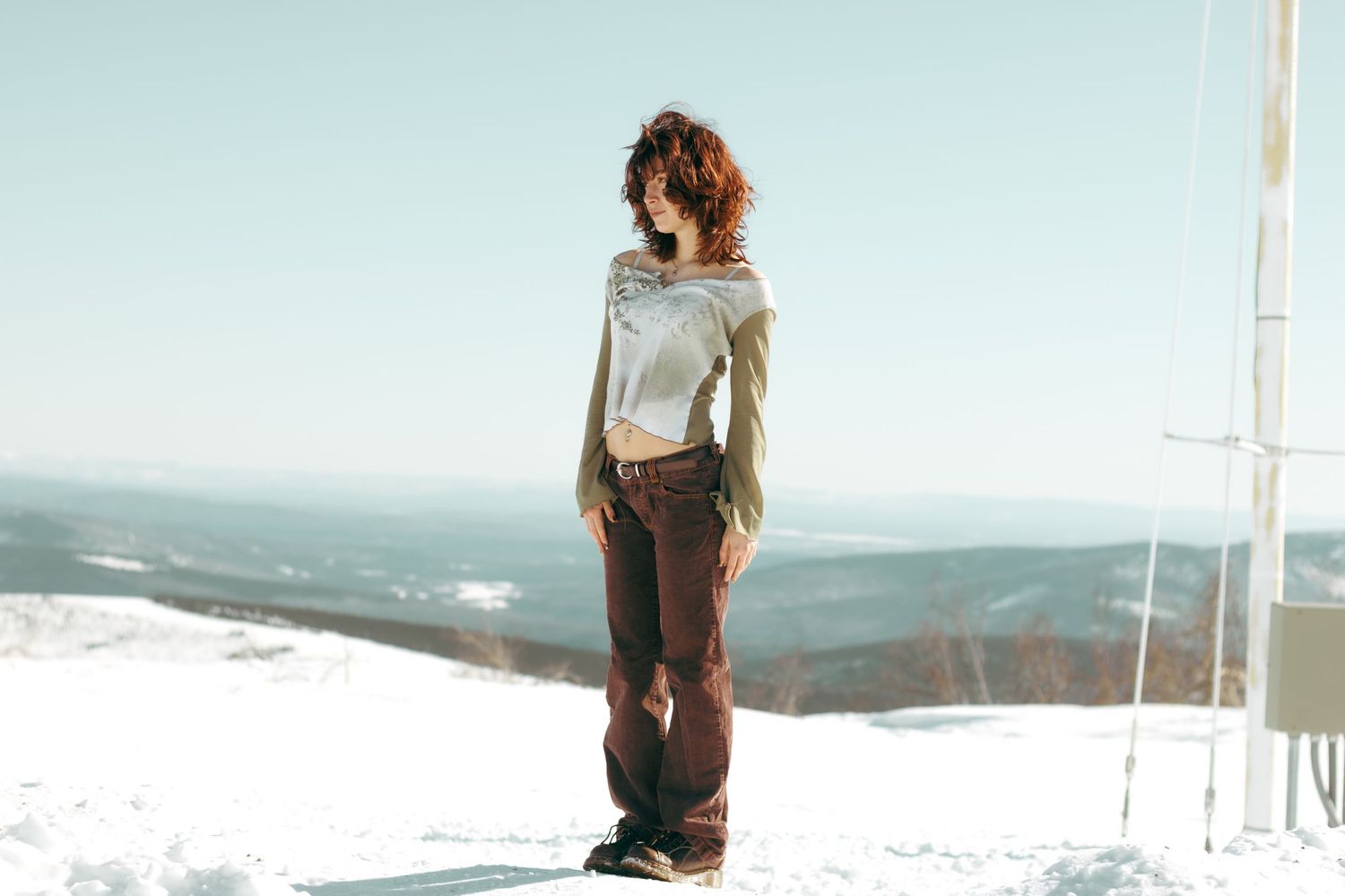 Person stands on snowy mountain peak with cloudy vista in background, wearing casual hiking attire.