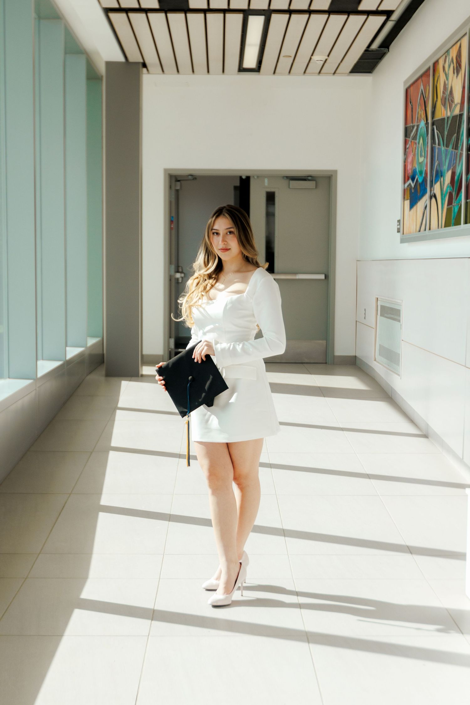 Young woman in white mini dress and heels holding black clutch in bright modern corridor.