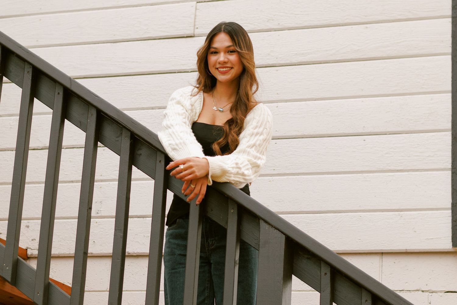 A person sits casually on outdoor wooden stairs while smiling warmly on a sunny day.