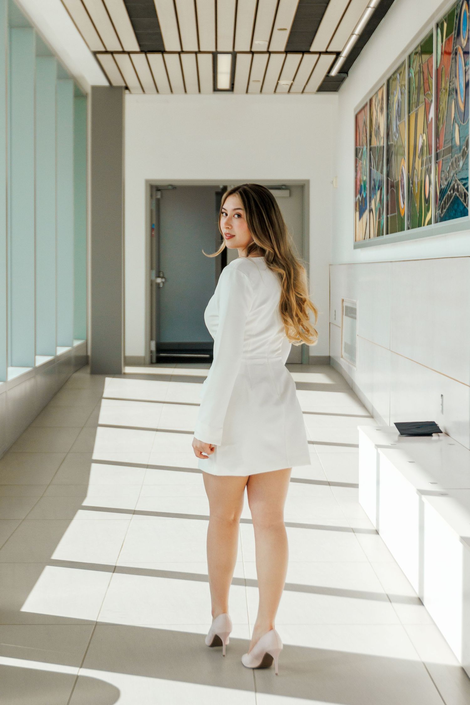 Woman in white mini dress and heels poses in a sunlit modern corridor with colorful artwork on the wall.