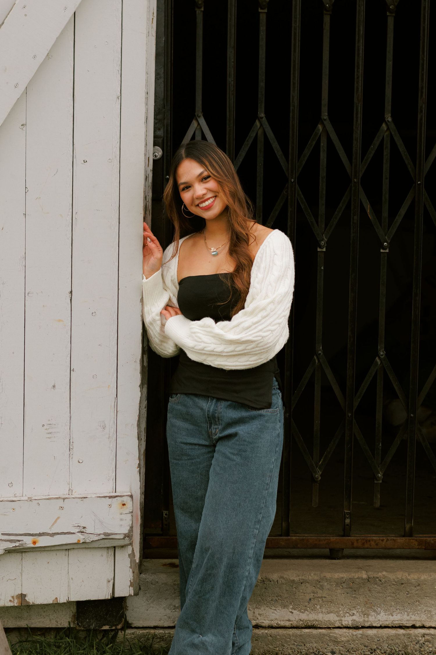 Casual outfit featuring a white cable knit sweater, black top and relaxed jeans posed in front of vintage metal gate.