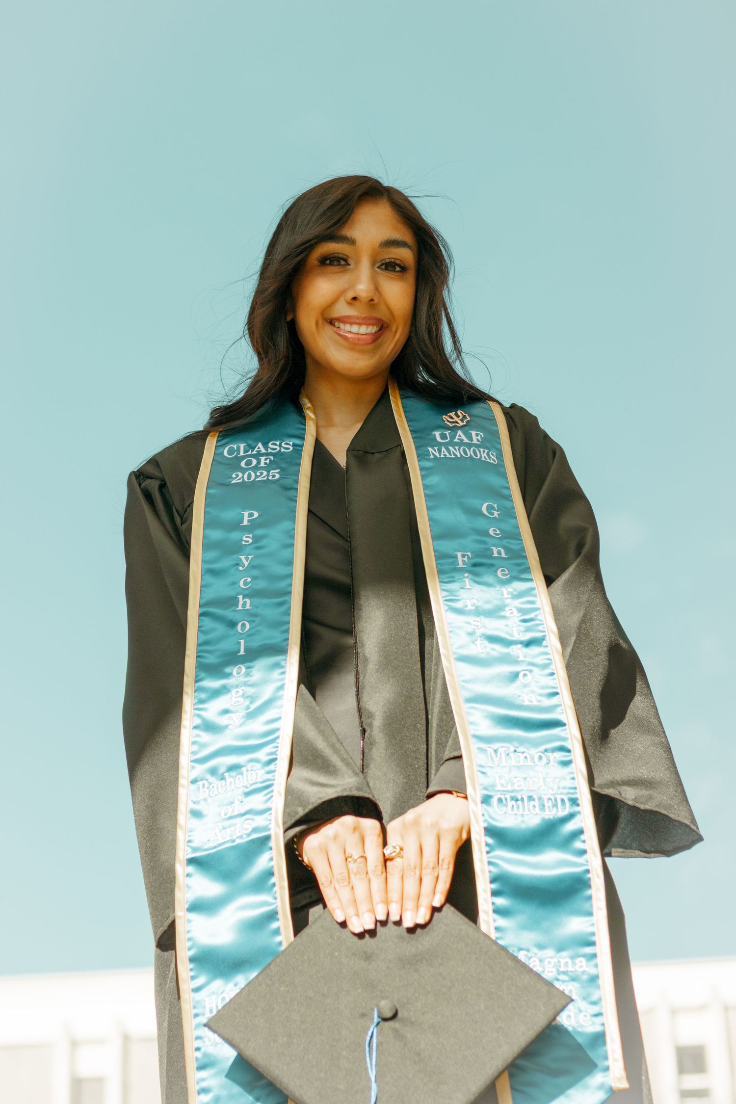 Graduate in black cap and gown wearing light blue graduation stoles smiles while posing for photos.