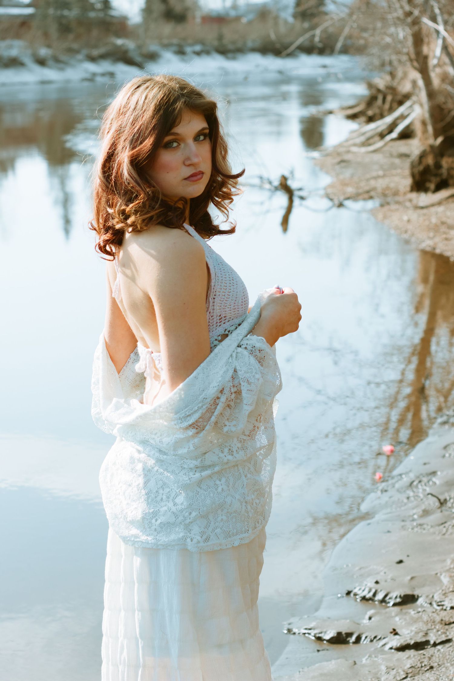 A dreamy portrait of a woman in a white lace dress standing by a serene river bank at sunset.