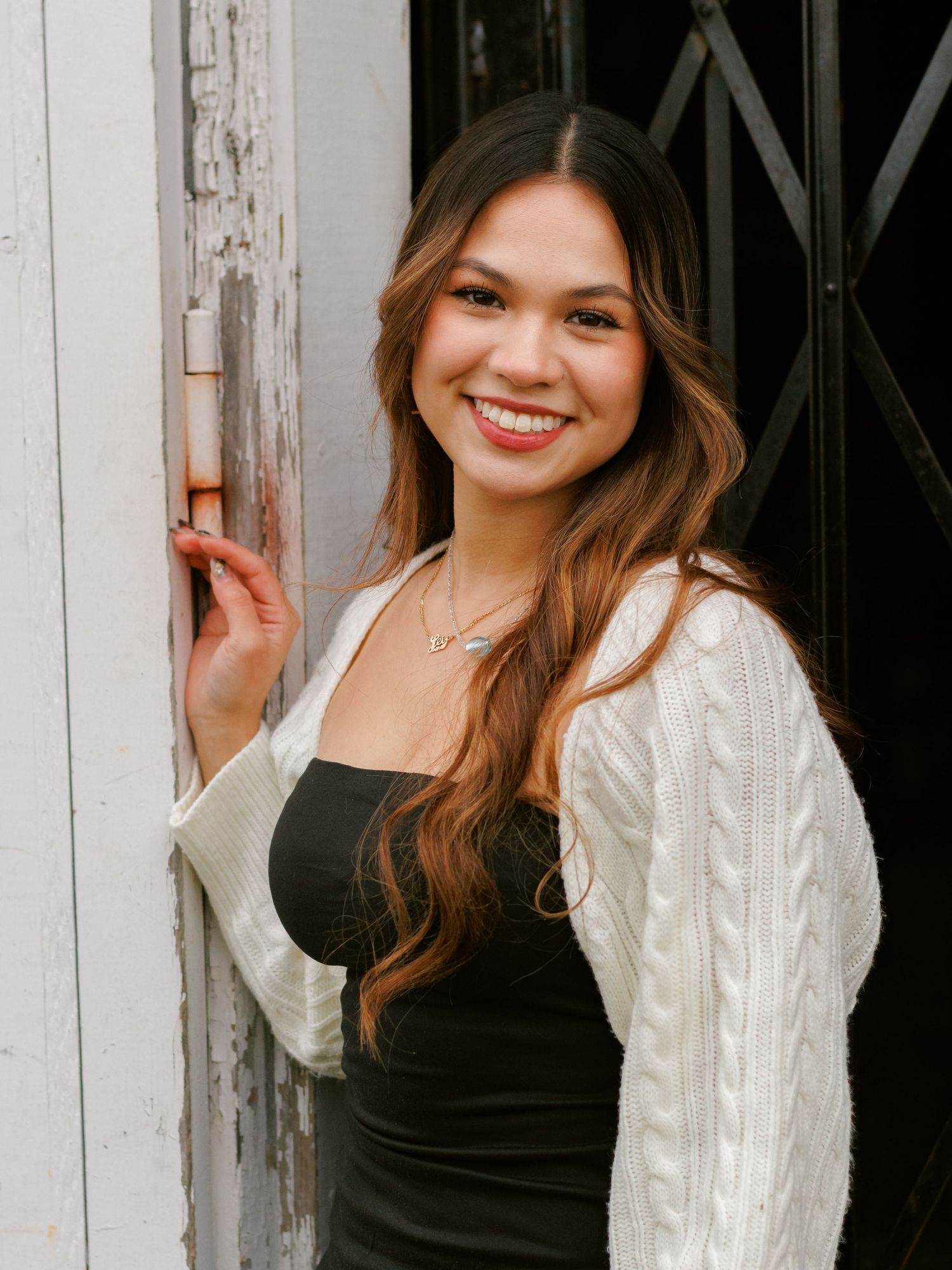 A person wearing a black tank top and white cable knit cardigan poses against a rustic white wooden wall.
