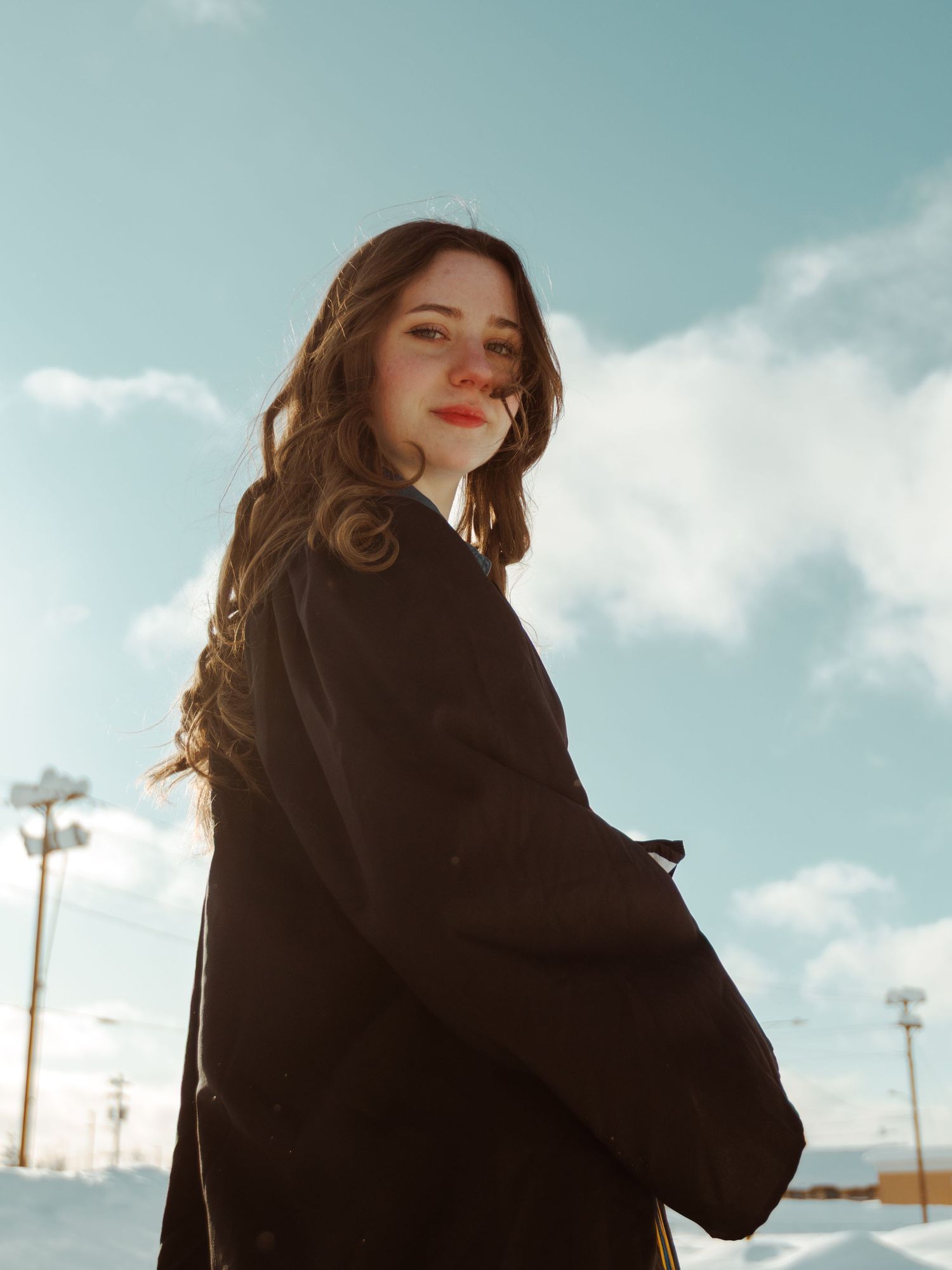 Figure in a black winter coat poses against a cloudy sky background in a stylish outdoor fashion photoshoot.