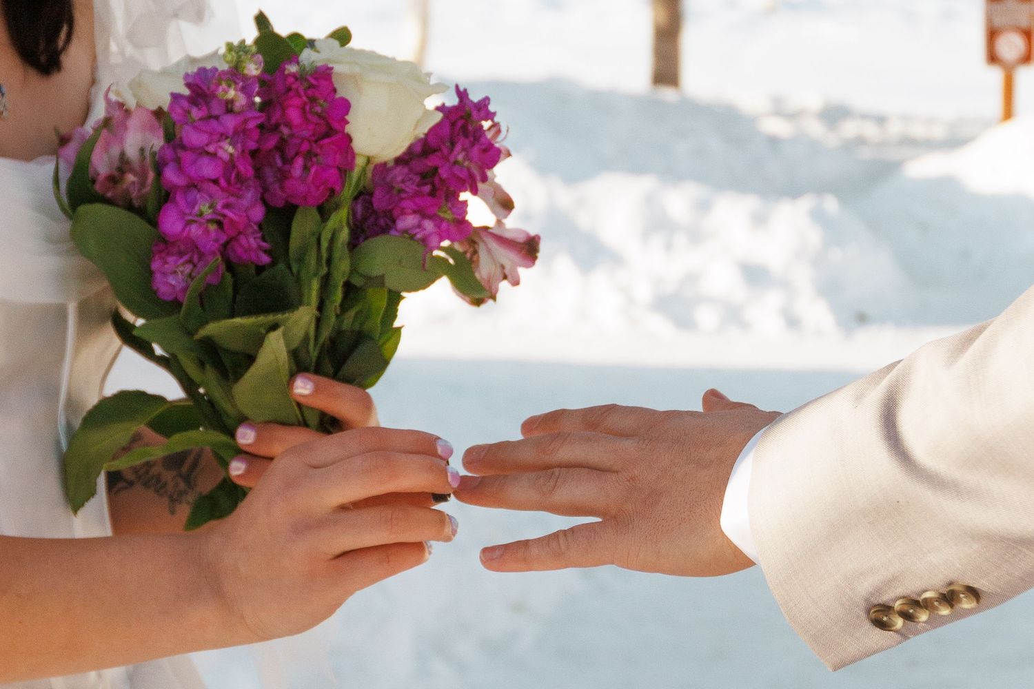 Bride holding purple flower bouquet while groom places wedding ring on her finger in snowy winter setting.