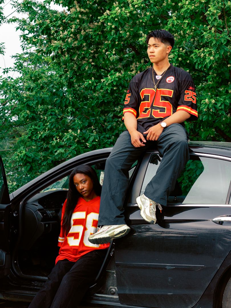 A person in a sports jersey numbered 25 sits on a black car.