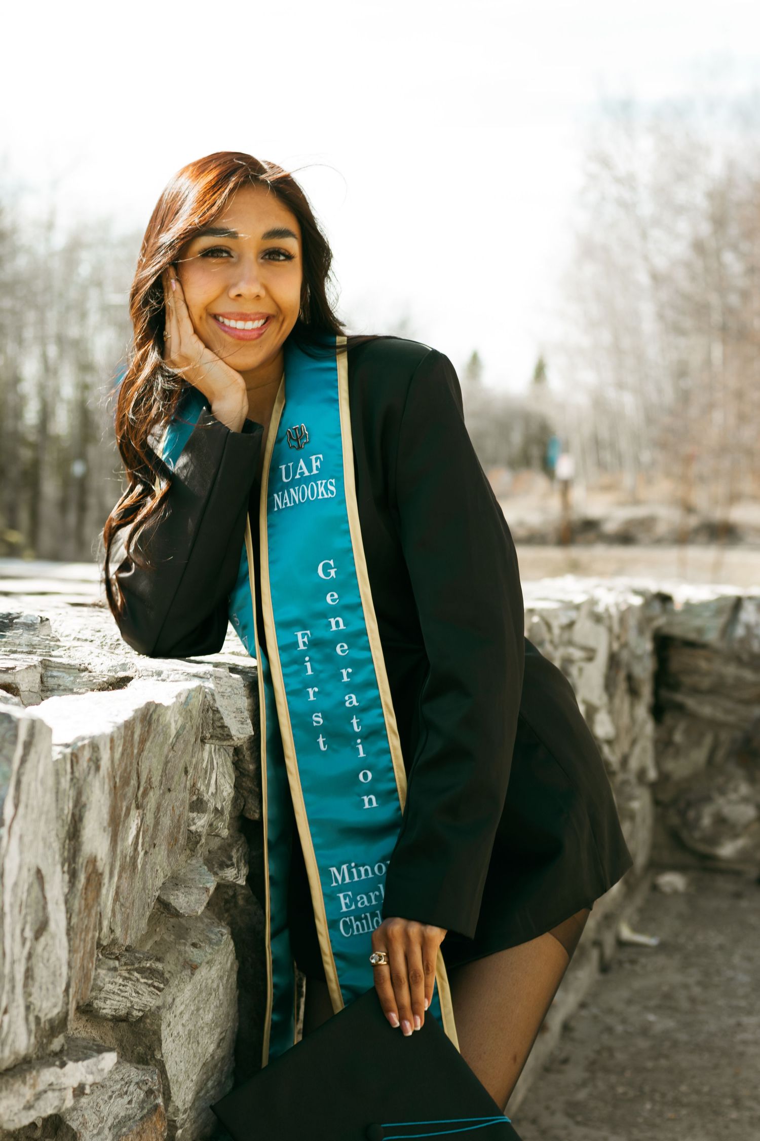 Person in black attire poses with a blue graduation stole against a stone wall backdrop in winter.