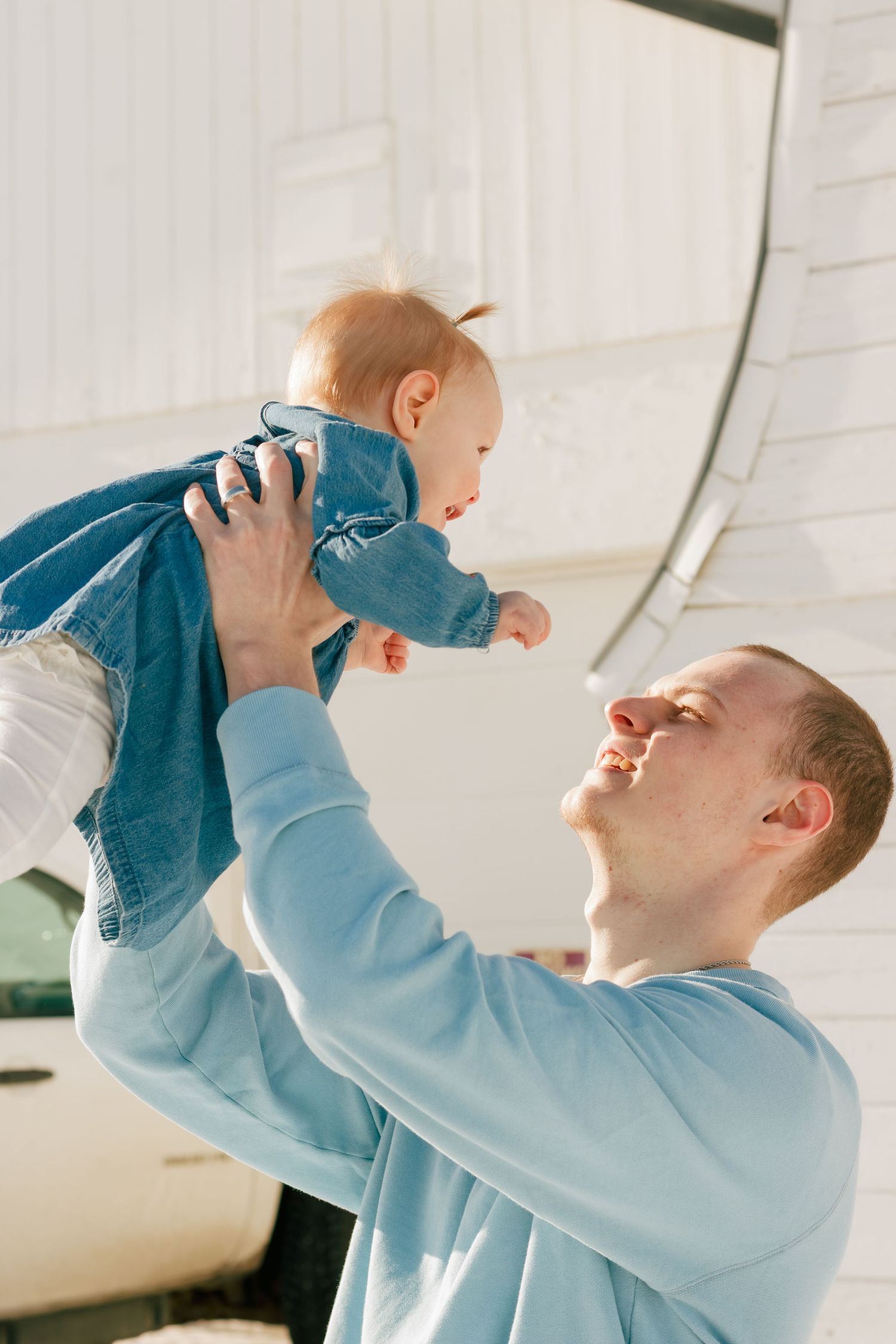 Parent playfully lifting baby in the air against white interior background.