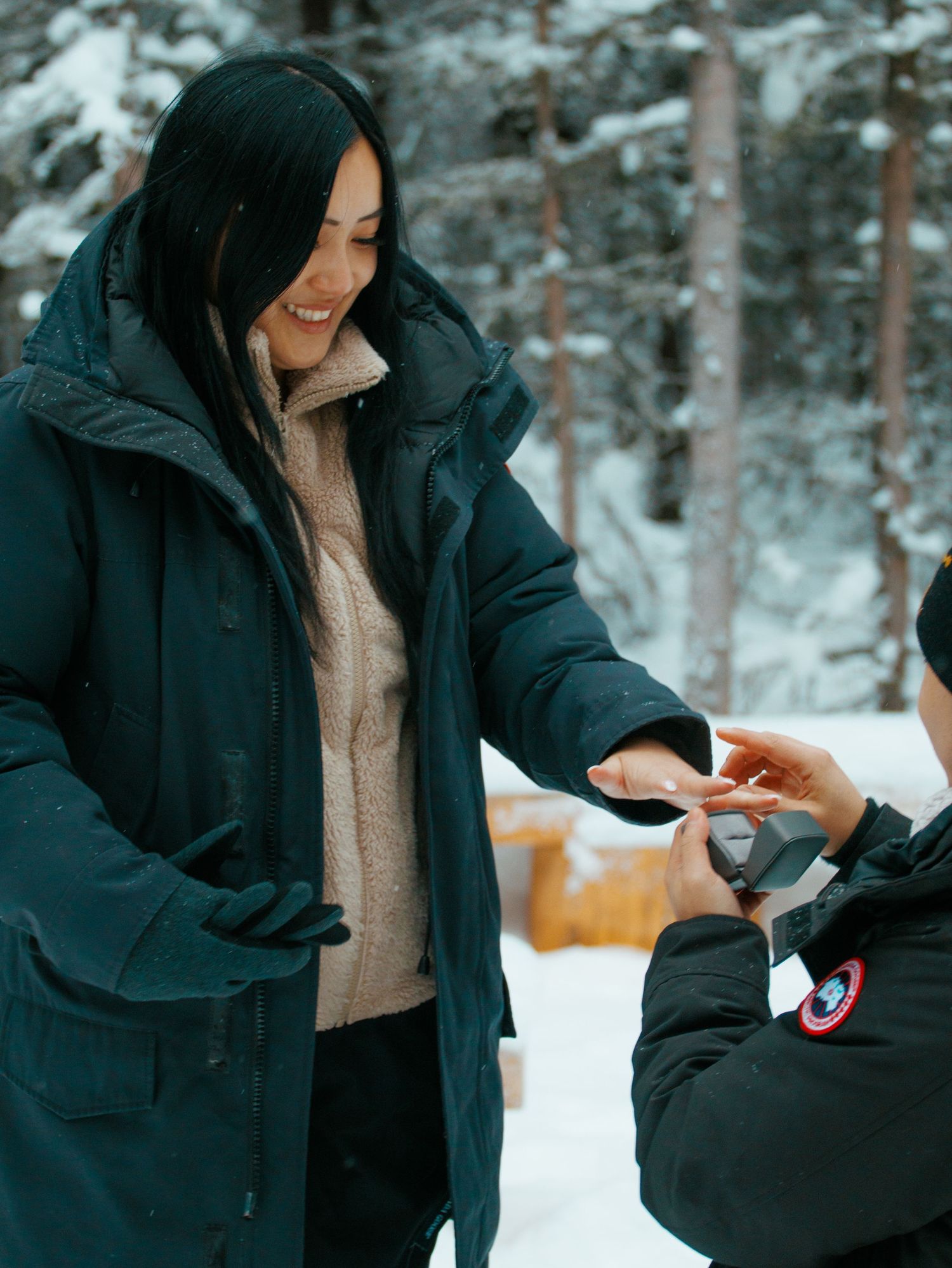 A couple in winter coats exchange a small object while standing in a snowy forest setting.