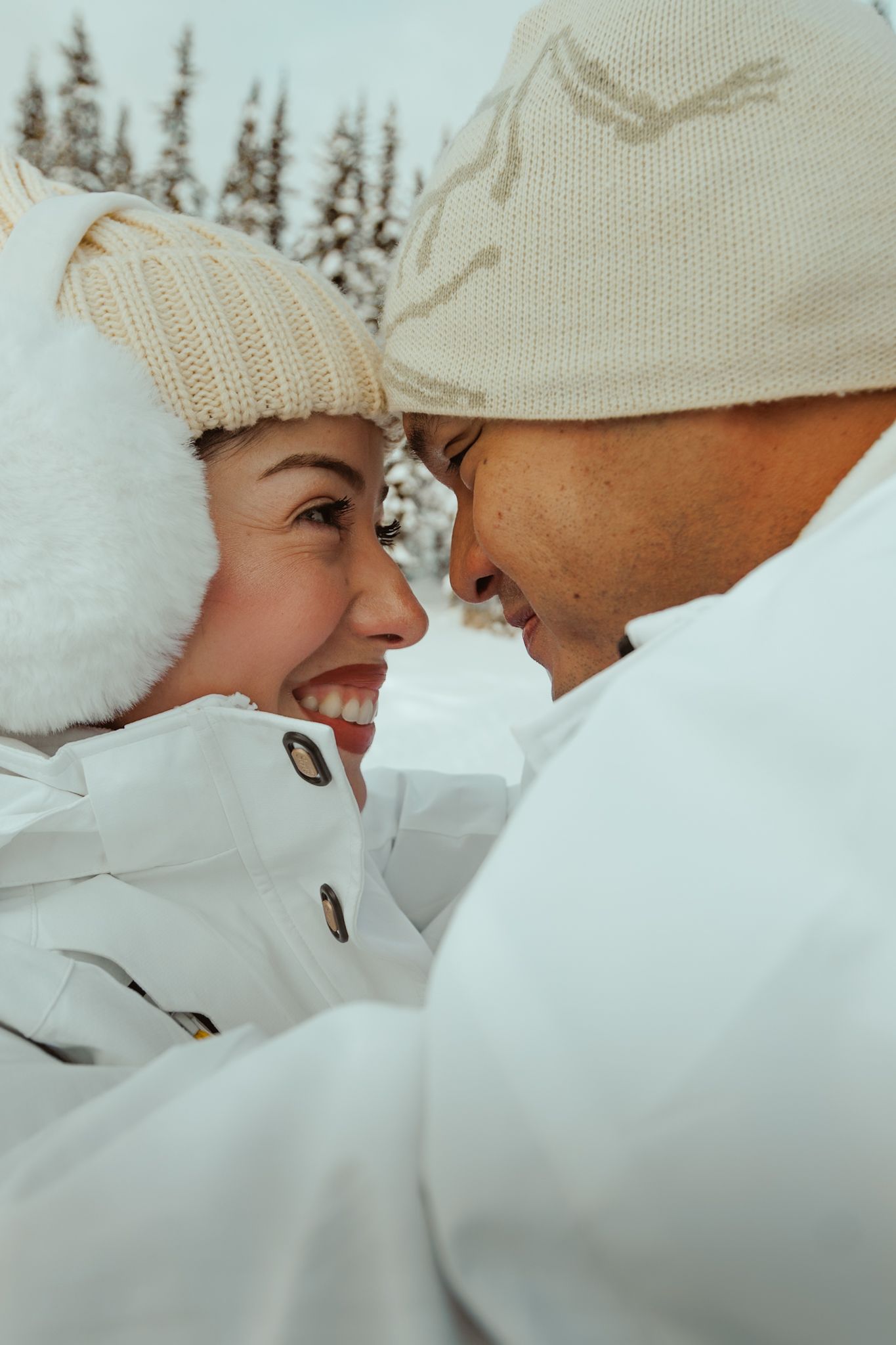 A close intimate winter moment between a couple wearing white coats and knit hats in a snowy setting.