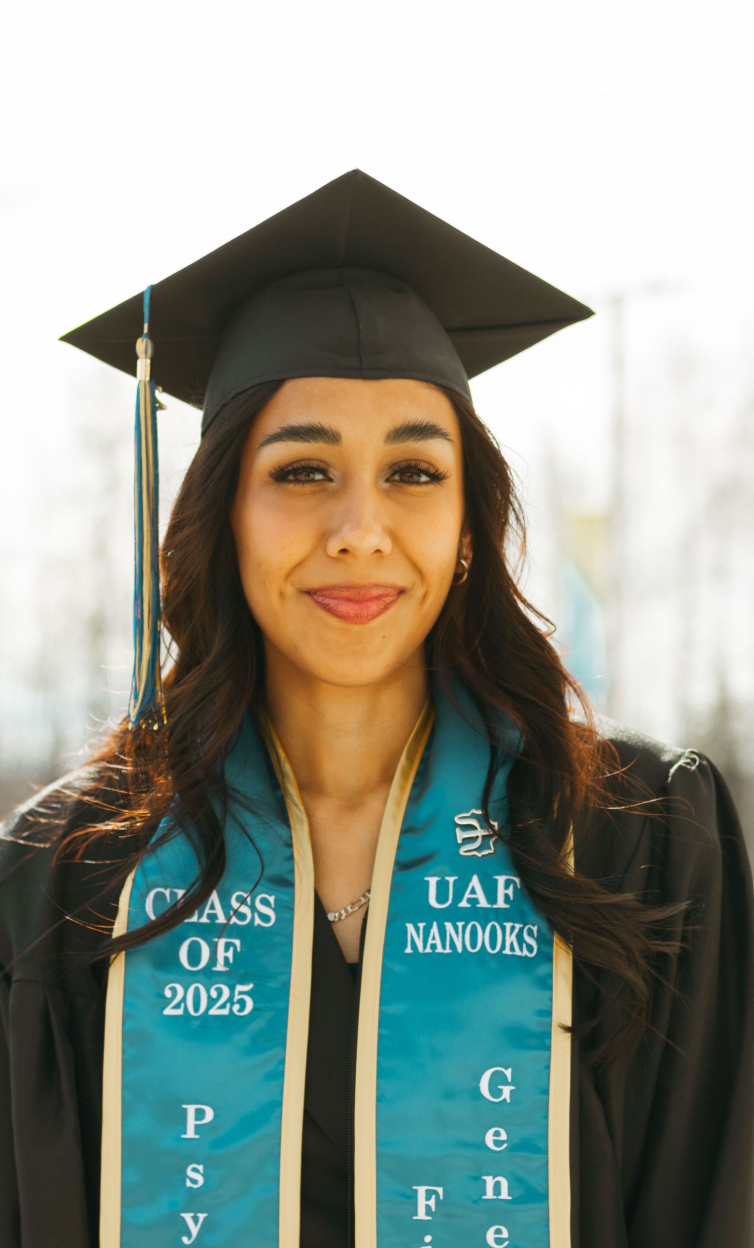 Graduate wearing black cap and gown with turquoise sash smiles in front of bright background.