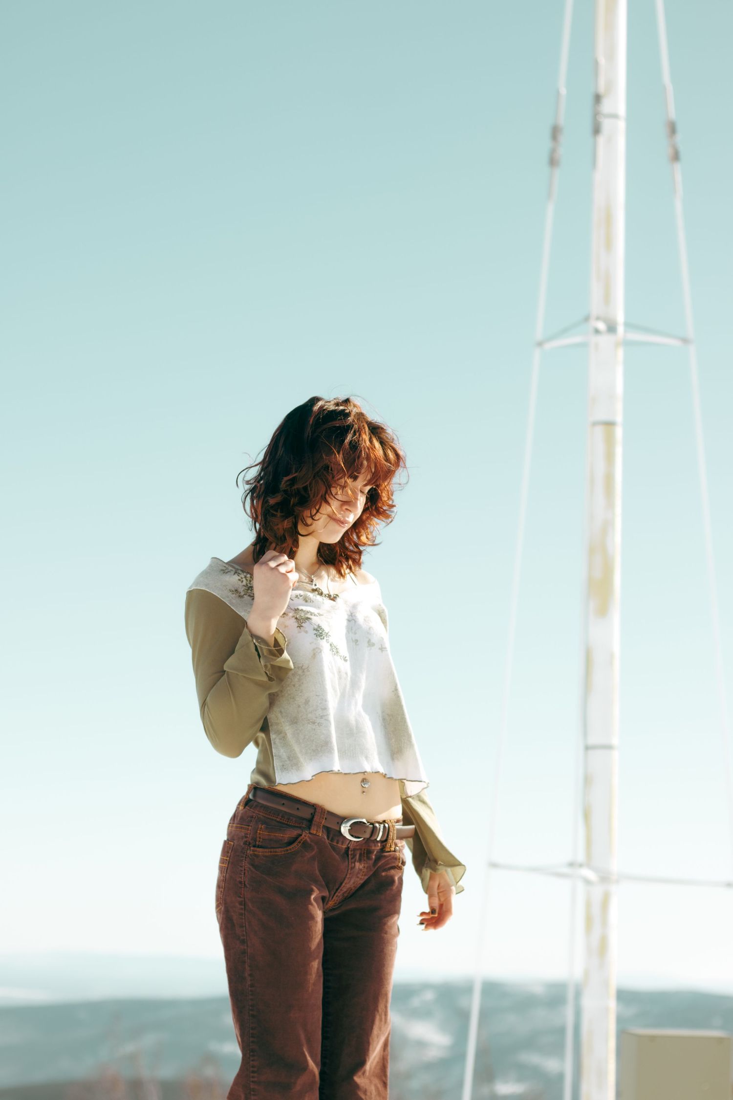 A person in a white cropped shirt and brown pants poses against a bright sky with utility poles in the background.