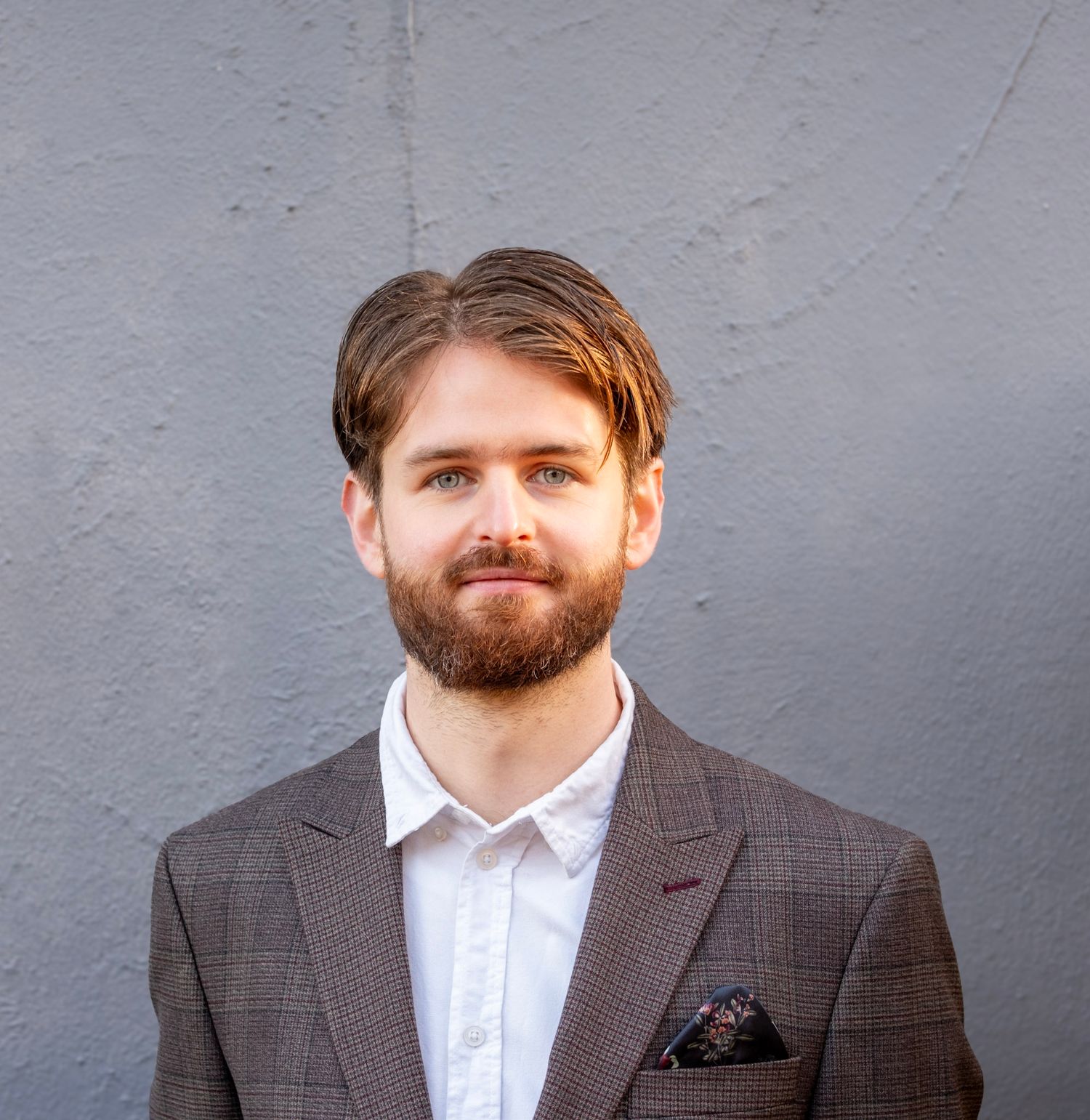 Young professional man with beard in gray blazer and white shirt posing against gray wall.