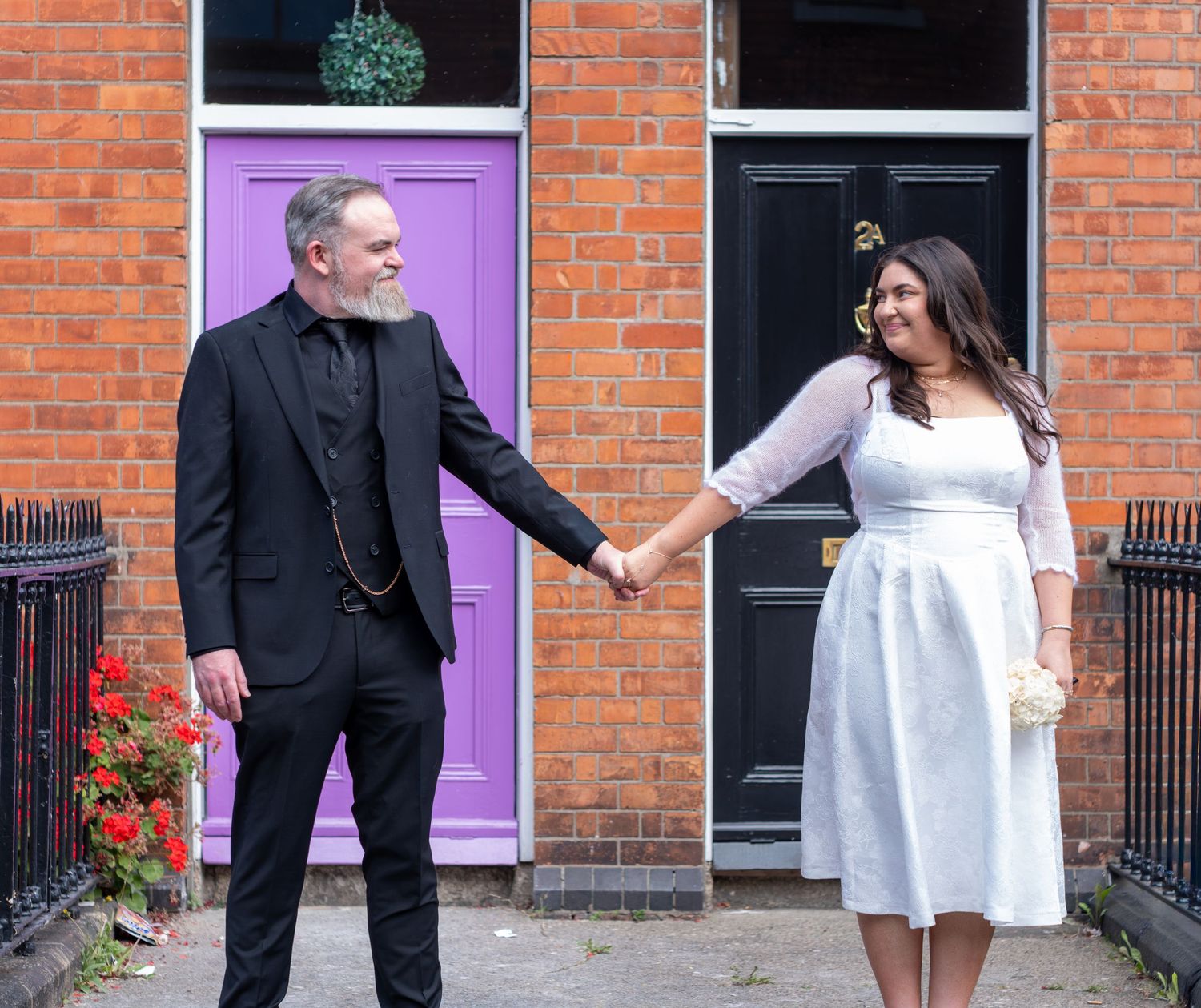 Couple in wedding attire holding hands outside colorful doors on a brick building street.