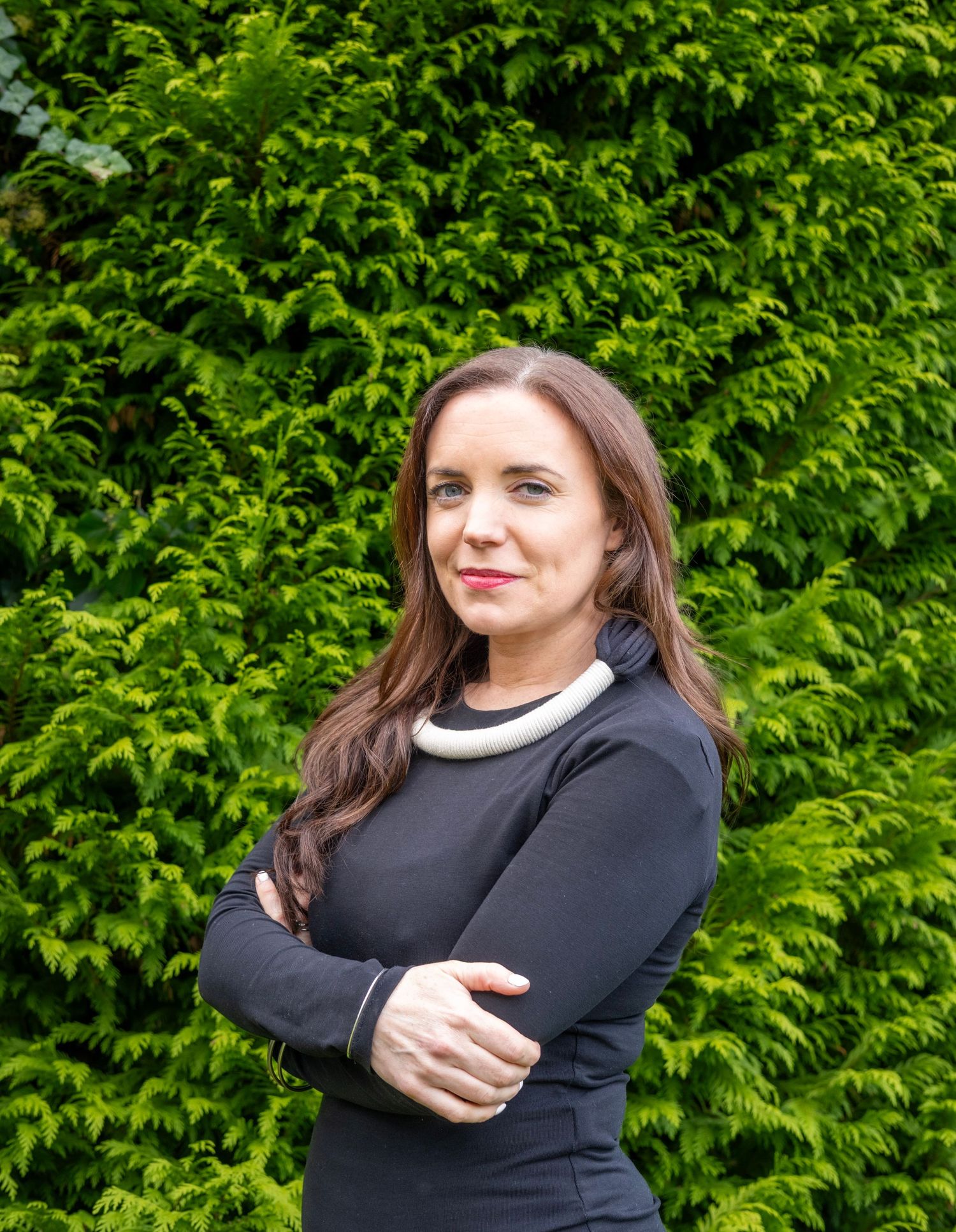 Professional woman in navy top with arms crossed stands before lush green hedge backdrop.