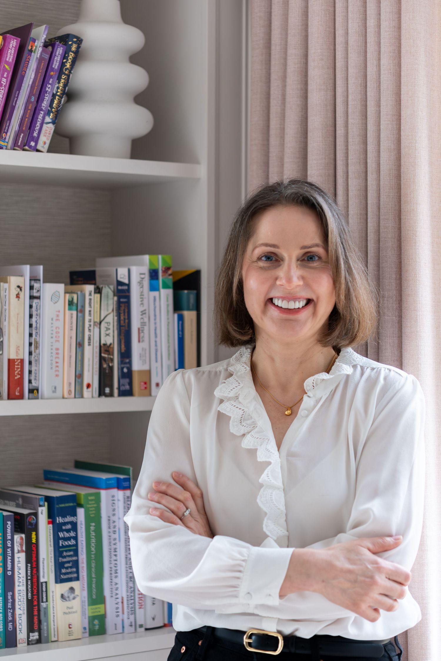 Smiling woman in white blouse stands arms crossed before bookshelf in bright, modern home office setting.