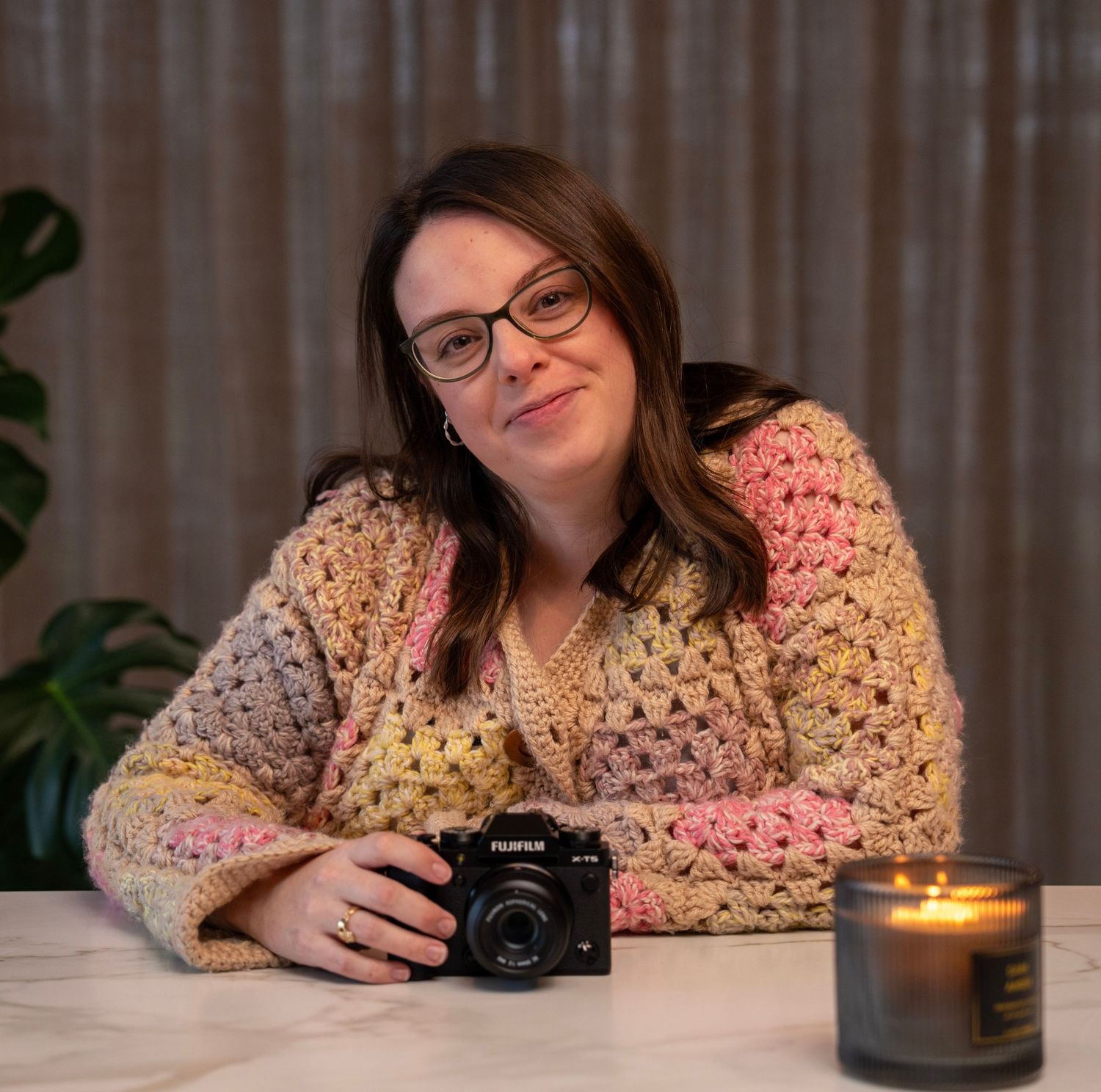 Photographer with camera sits at table with cozy sweater and lit candle in warm indoor setting.