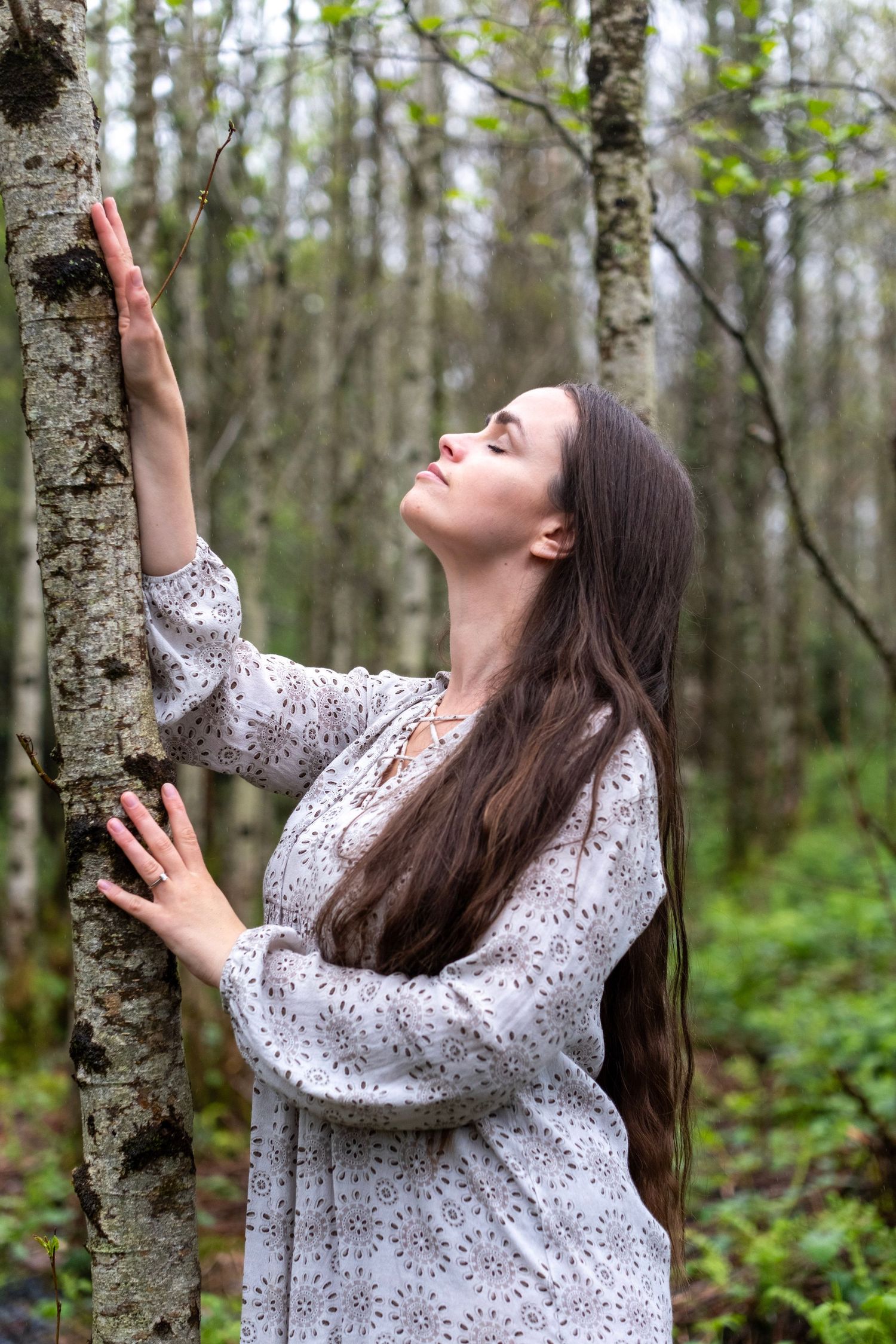 A person in a white floral dress leans against a birch tree trunk in a peaceful forest setting.