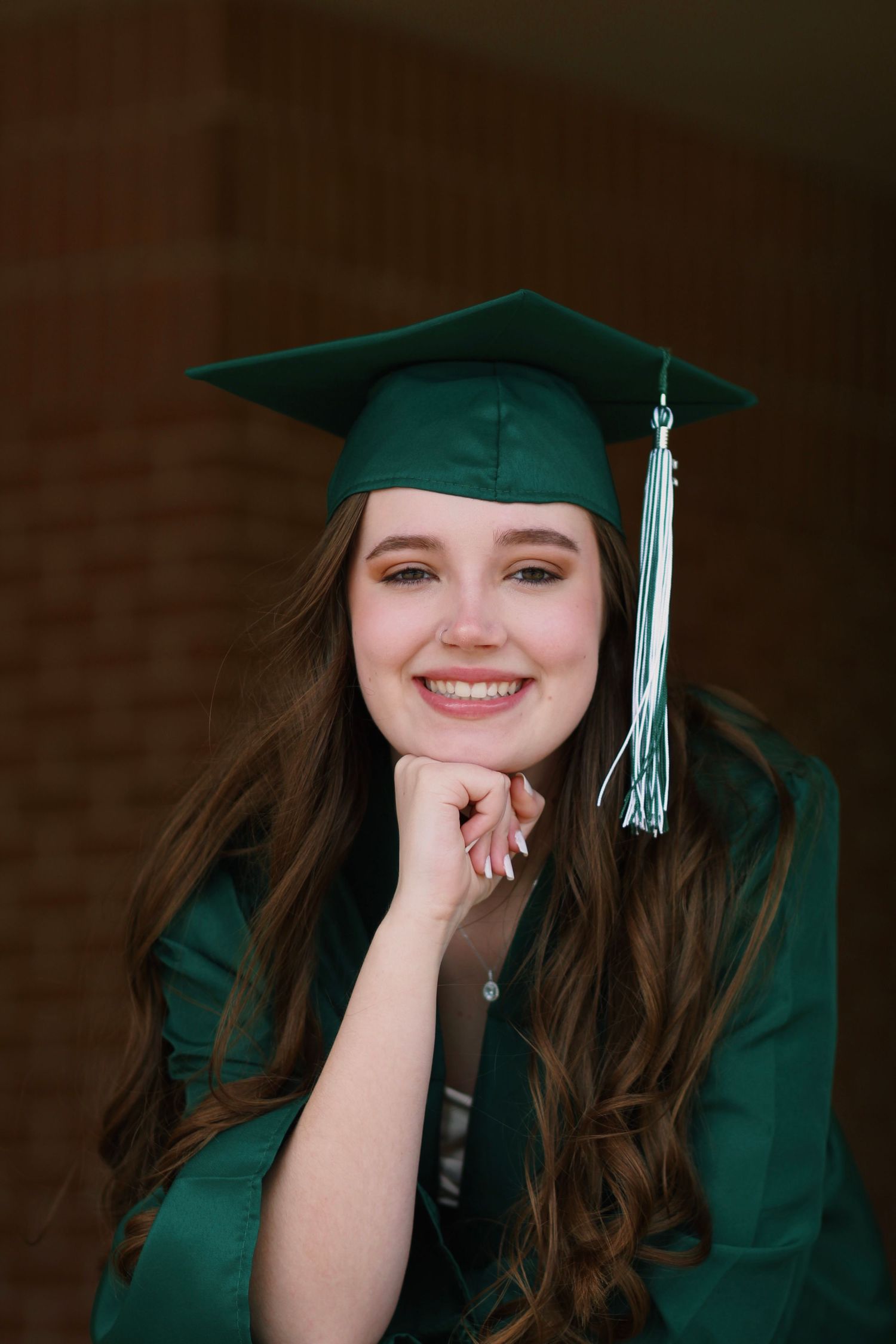 A graduate in emerald cap and gown poses with hand on chin against brick background.