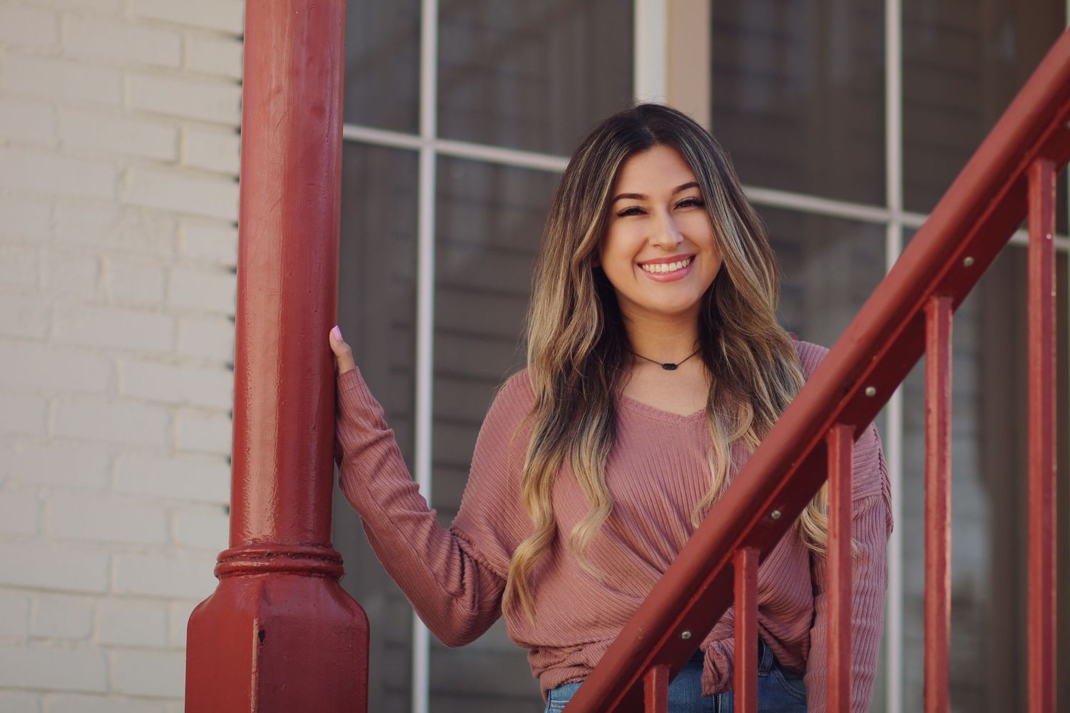 Portrait by red railing wearing a pink sweater against white brick wall.