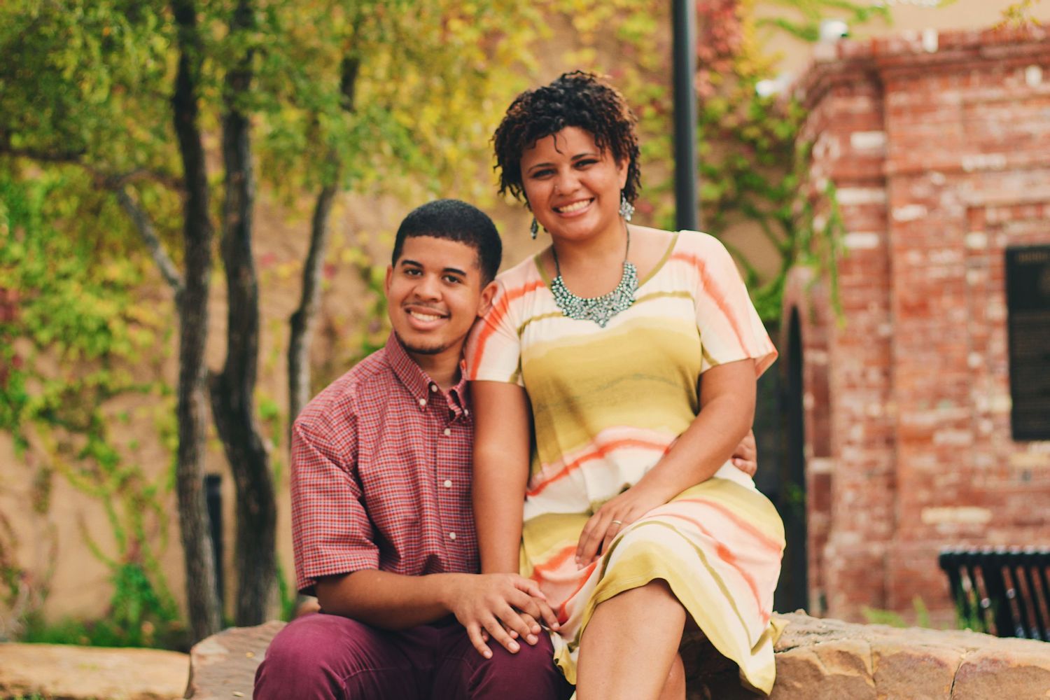 A couple sits together outdoors wearing colorful casual attire against a natural brick and foliage backdrop.