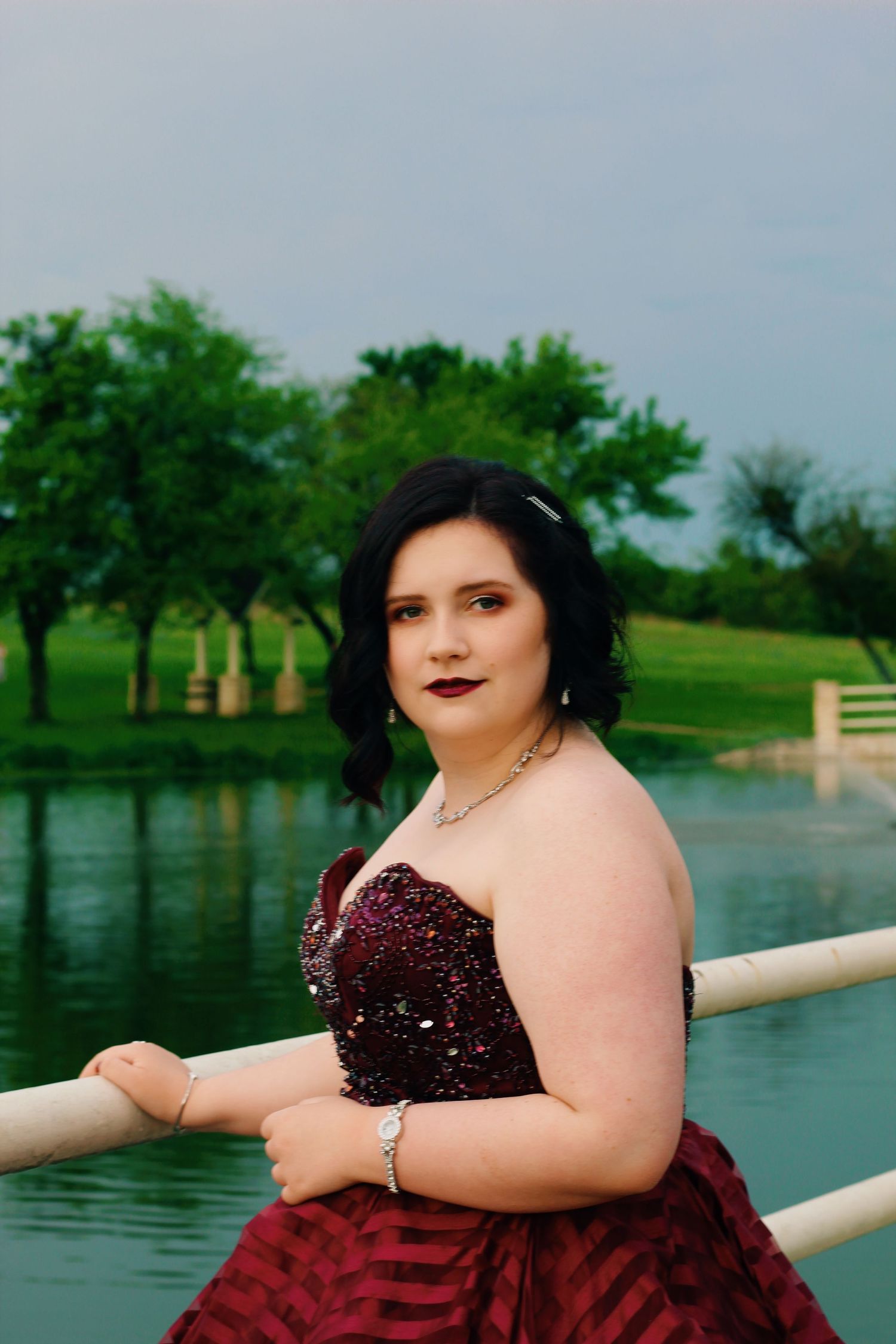 A portrait by a lake with trees in the background featuring a burgundy sequined formal dress.