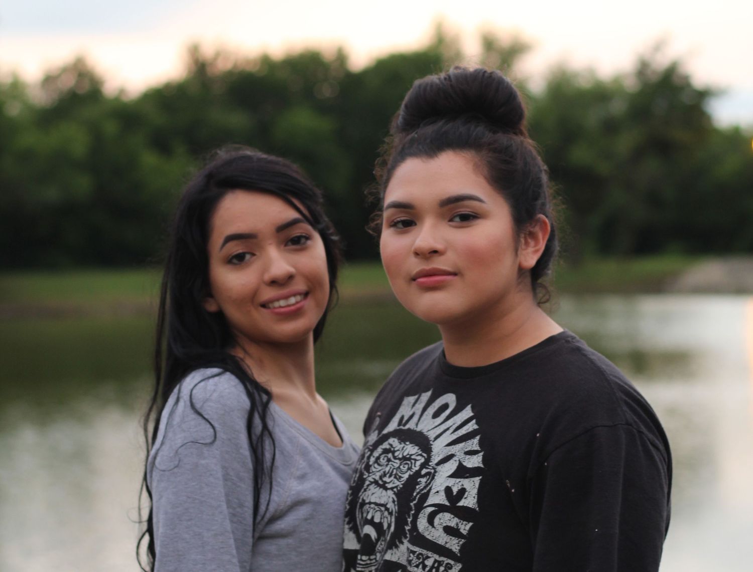 Two cousins in casual wear pose together near water at dusk.