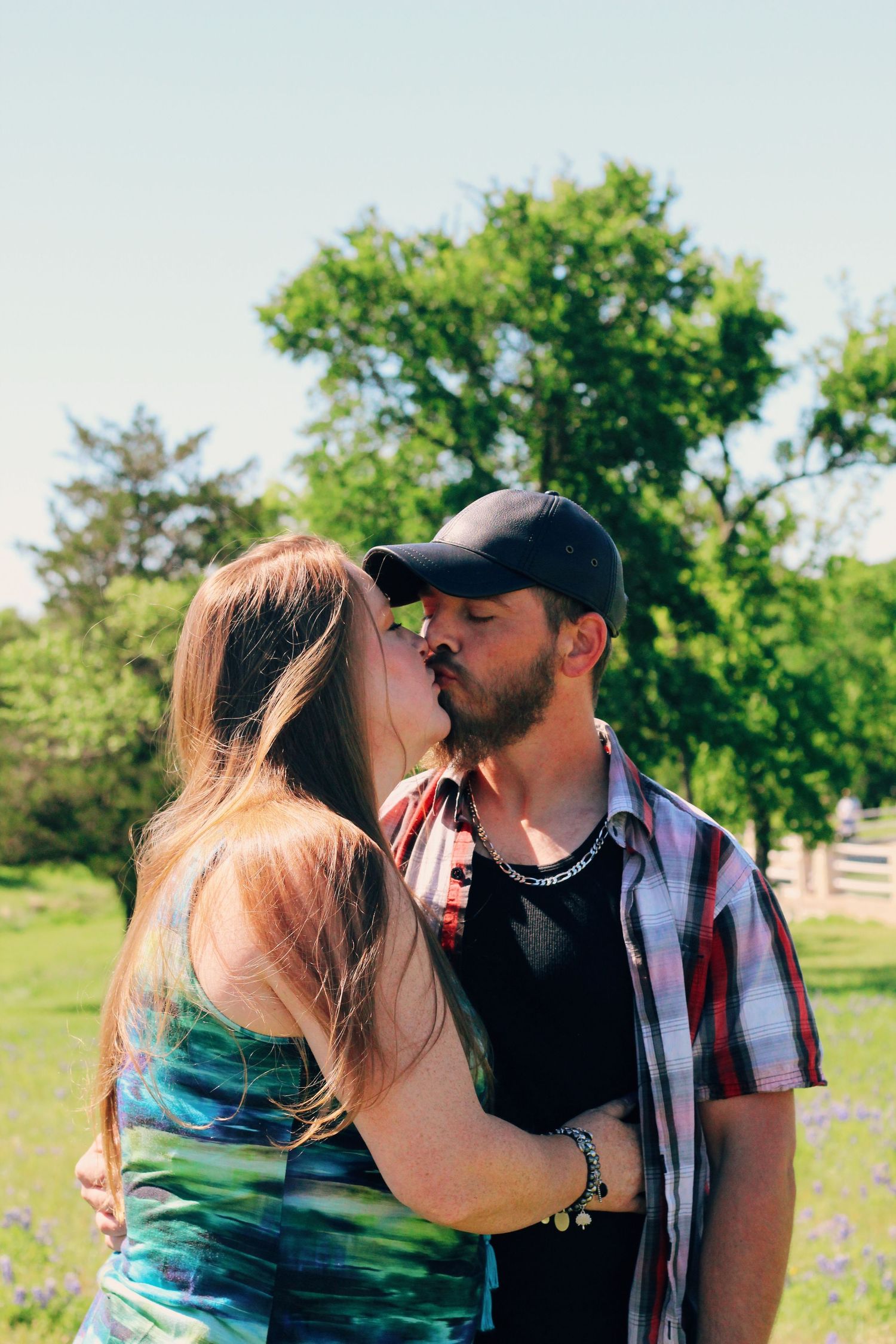 A couple shares a kiss outdoors with green trees and a fence in the background.