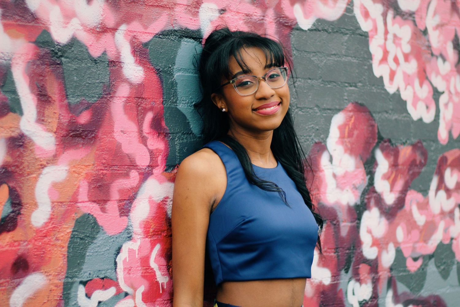 A person in a blue crop top poses against a vibrant pink and gray graffiti wall.
