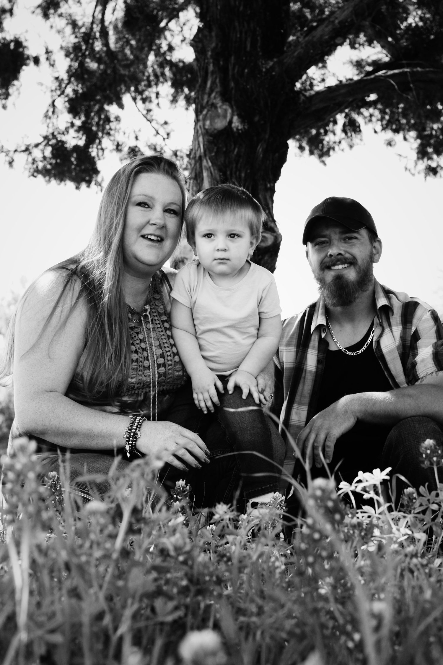 Black and white outdoor family portrait under large tree with wildflowers in foreground.