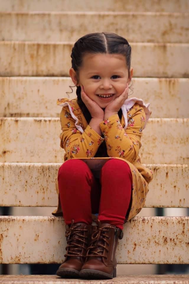 Young child in yellow jacket and red pants sitting on stone steps with hands on cheeks.