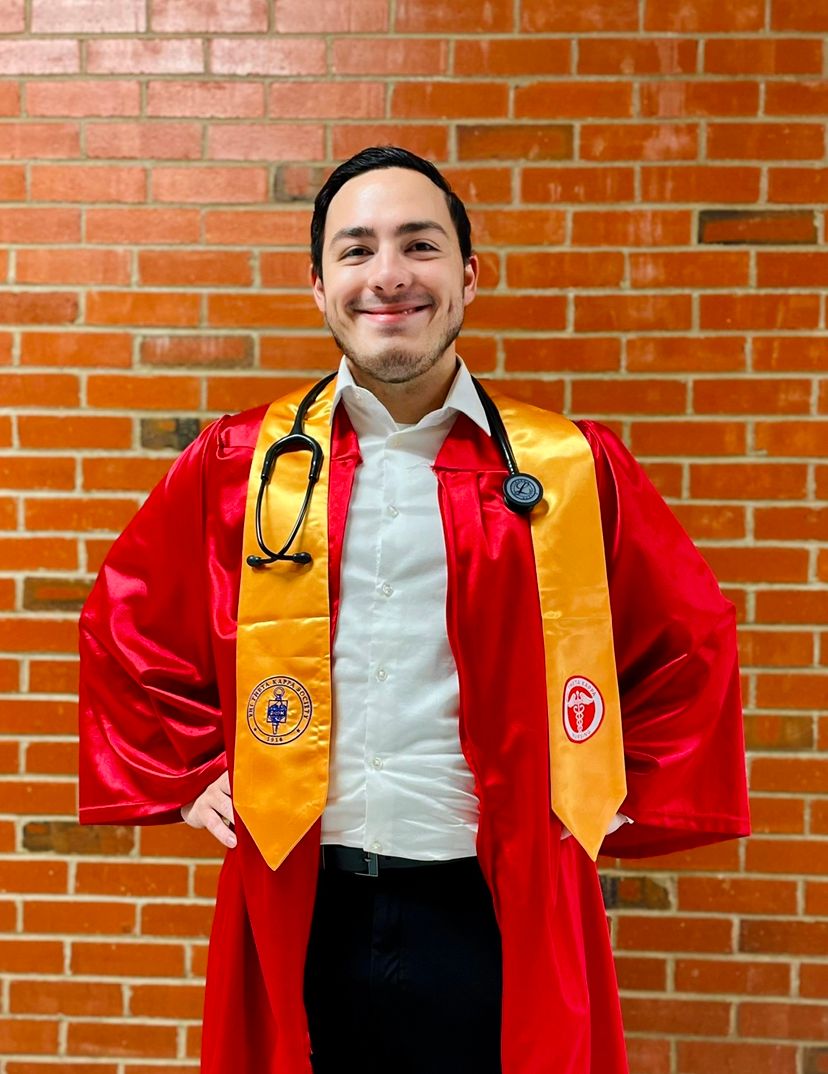 Graduate in red academic regalia and gold stoles with stethoscope against brick wall.