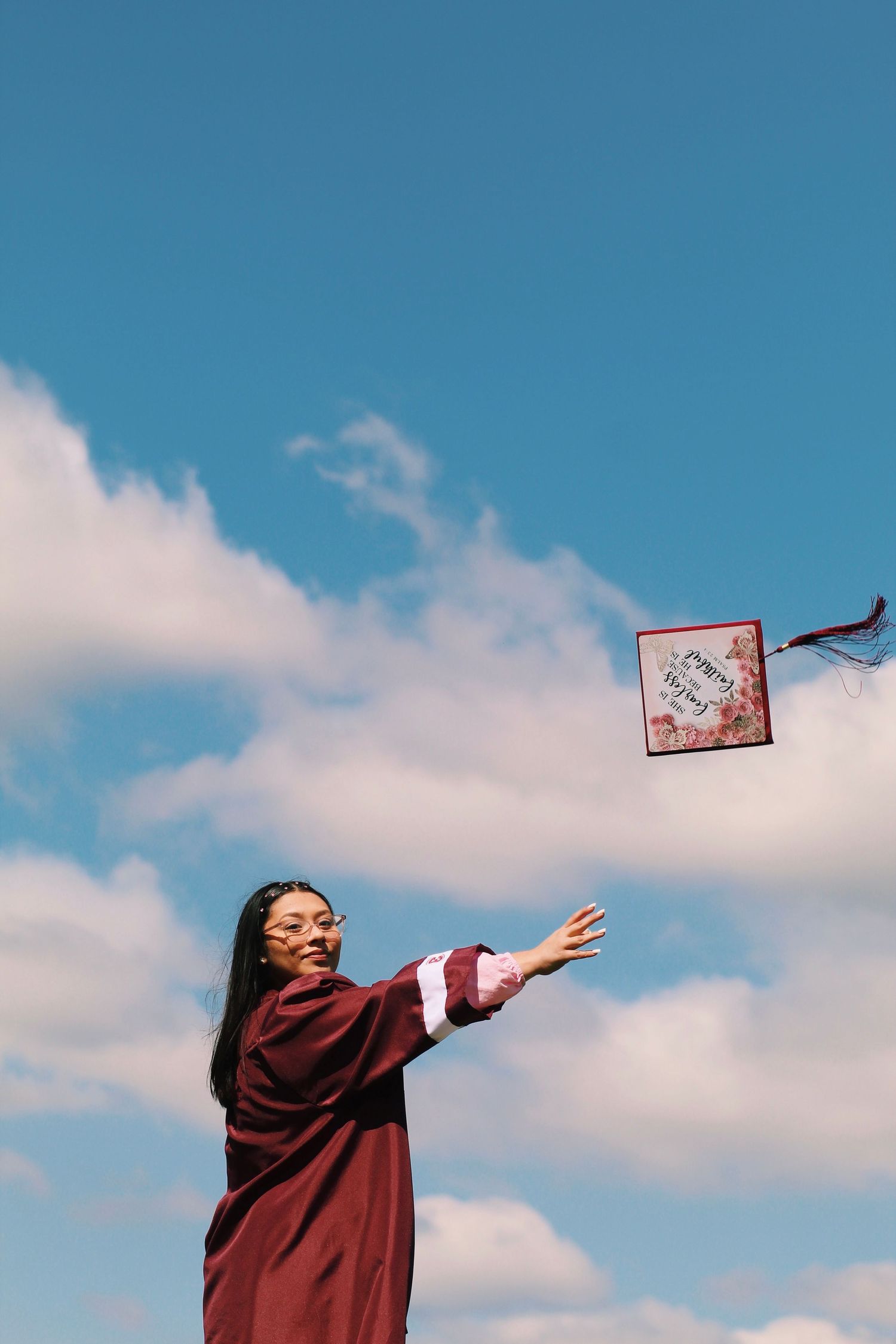 Graduate in maroon gown tossing cap against blue sky with clouds.