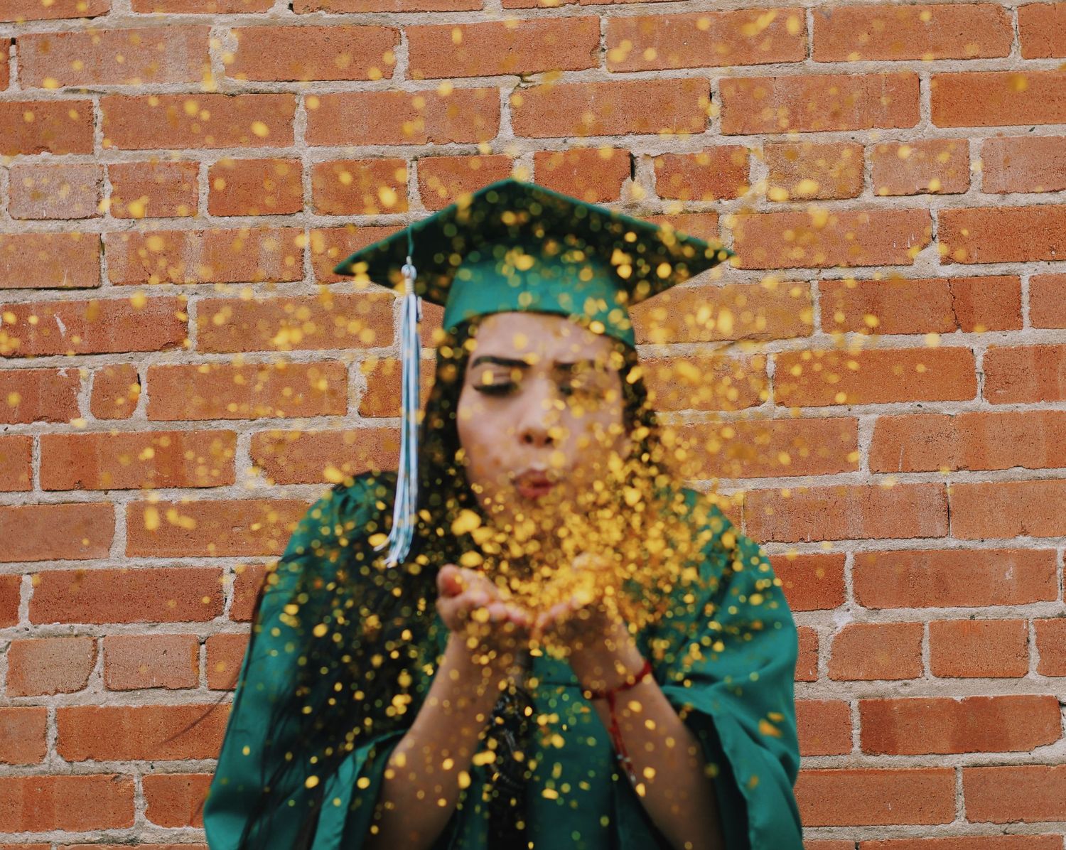 Graduate in green cap and gown blowing gold glitter against a brick wall.