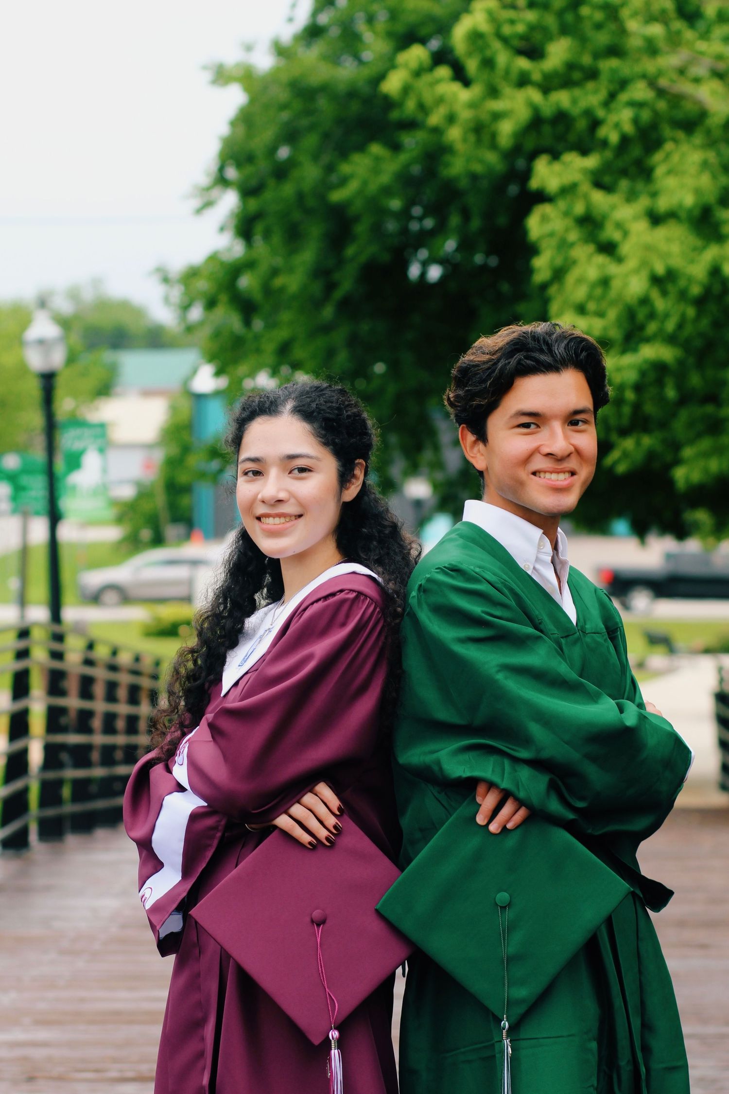 Two graduates in maroon and green gowns pose back-to-back with their diplomas.