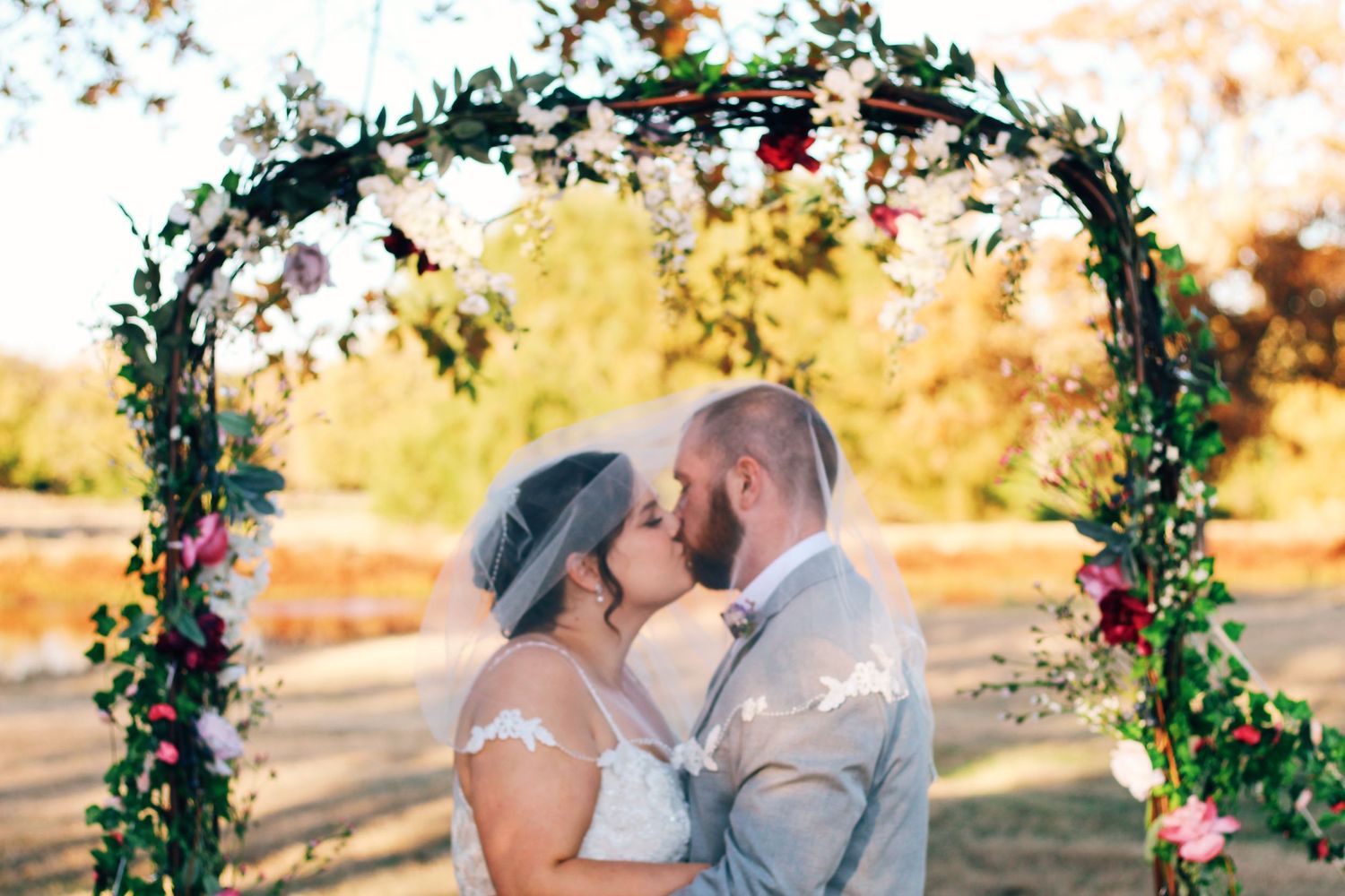Newlyweds kiss under a floral wedding arch during their outdoor ceremony.