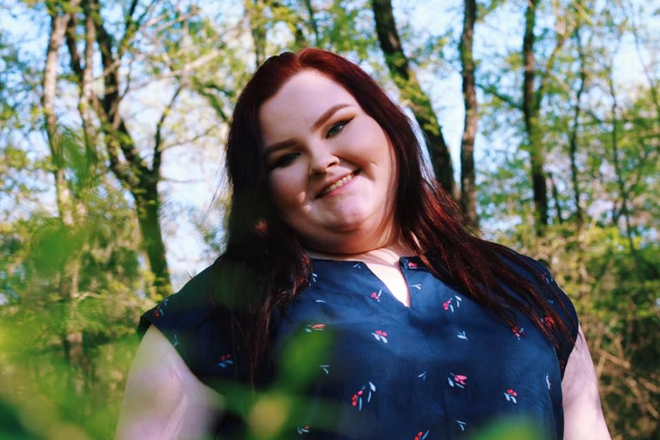 Natural outdoor portrait of a smiling figure in patterned blue top framed by green foliage.