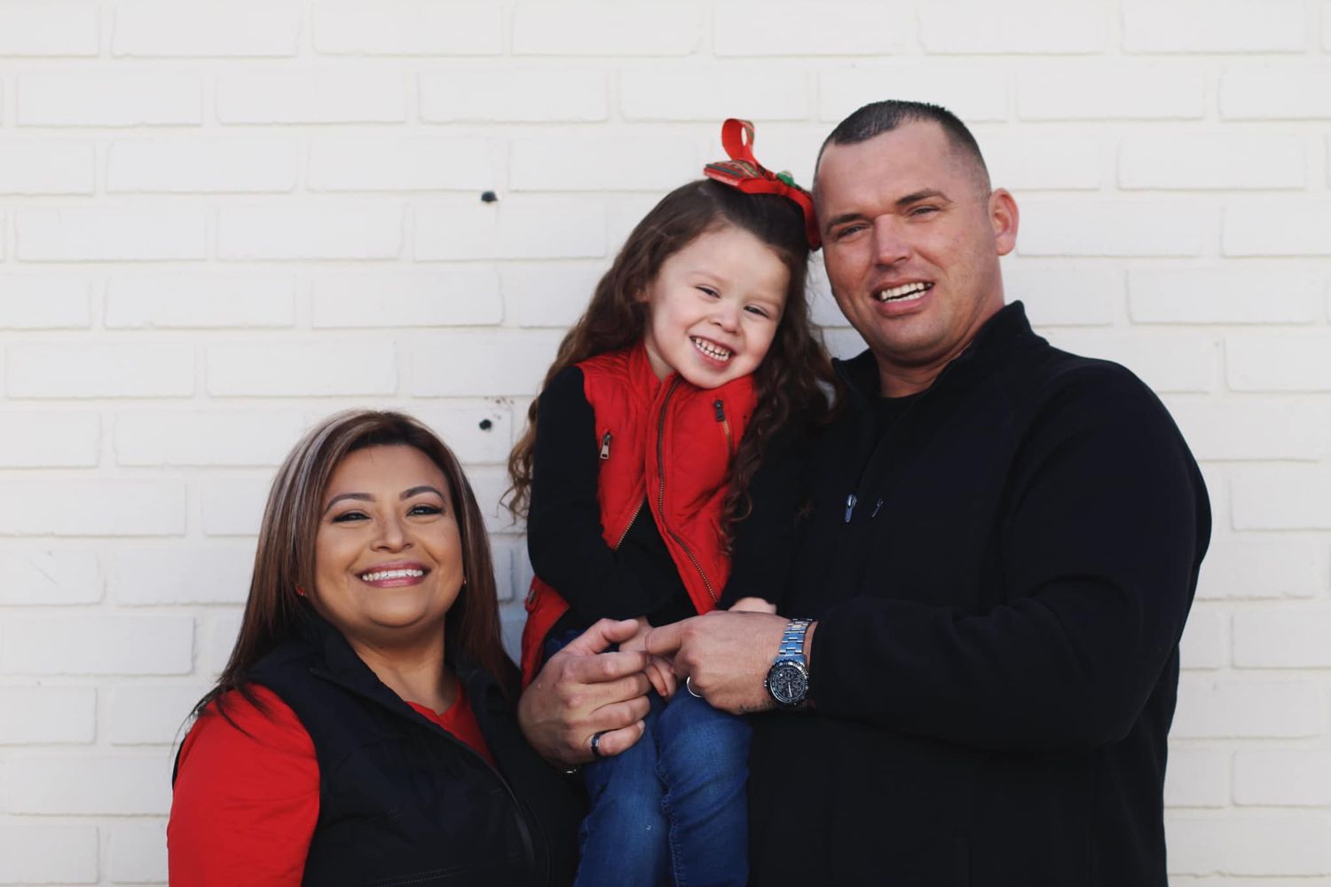 Family of three wearing red and black poses against white wall for holiday photo.