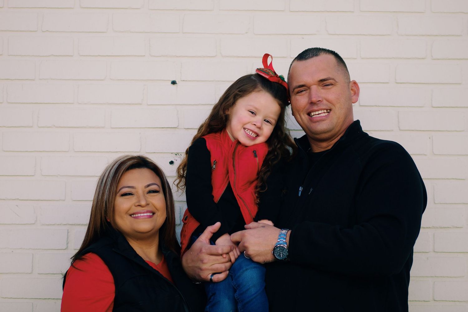 Family portrait with parents and young child wearing coordinating red and black outfits against light colored wall.