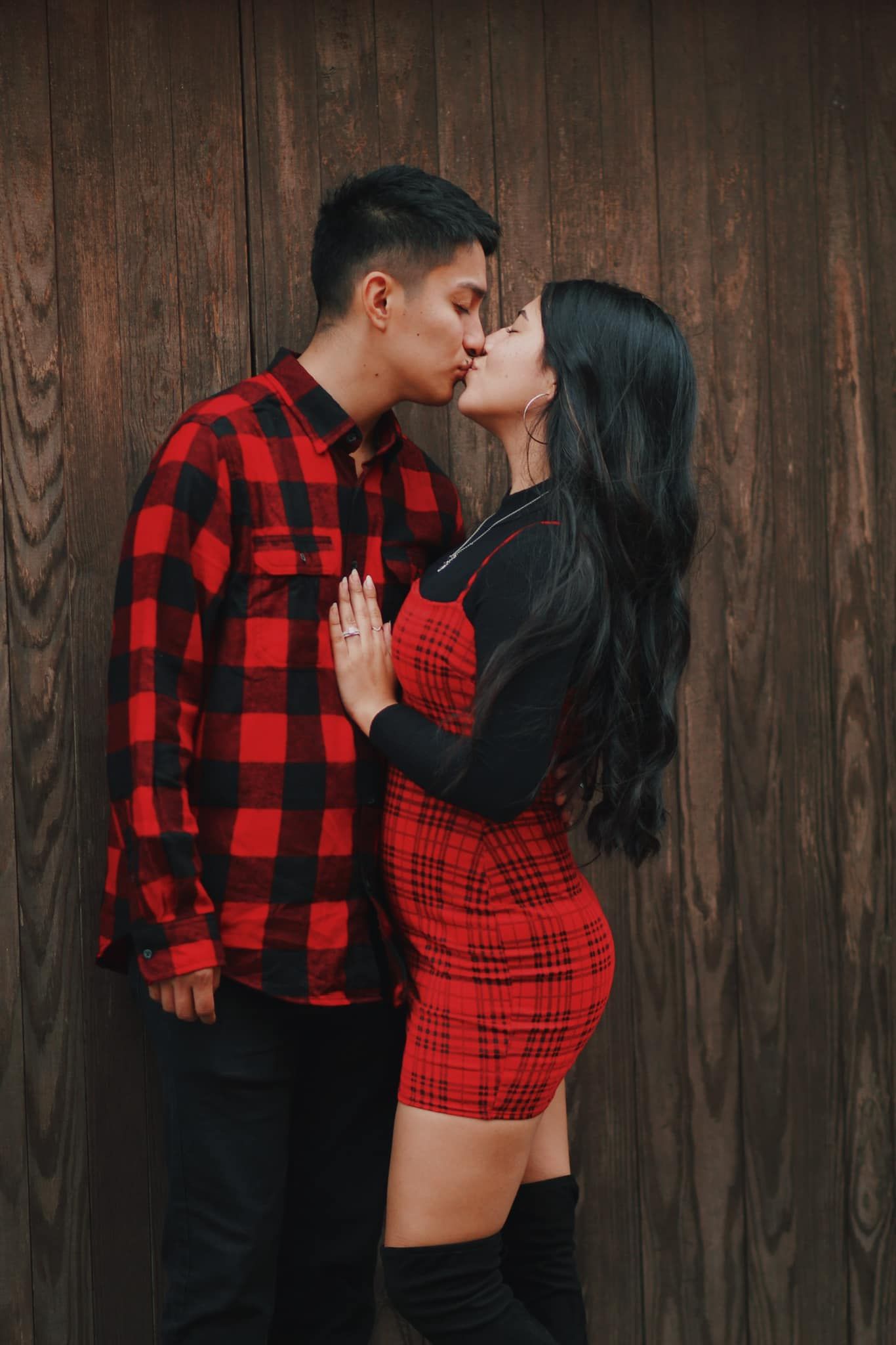 Romantic couple in matching red plaid outfits sharing an intimate moment against wooden wall.